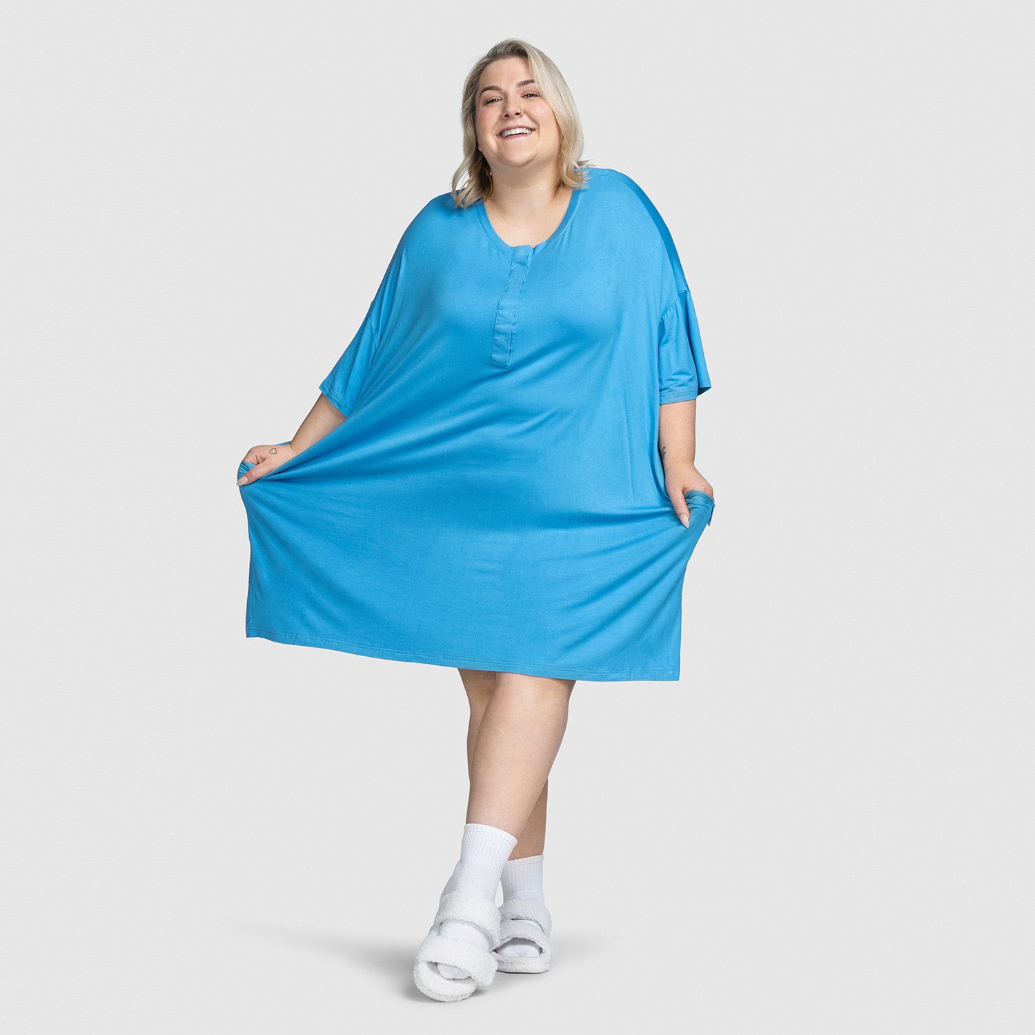Smiling woman wearing a loose blue dress, holding the sides of the dress, with white socks and fuzzy slippers.