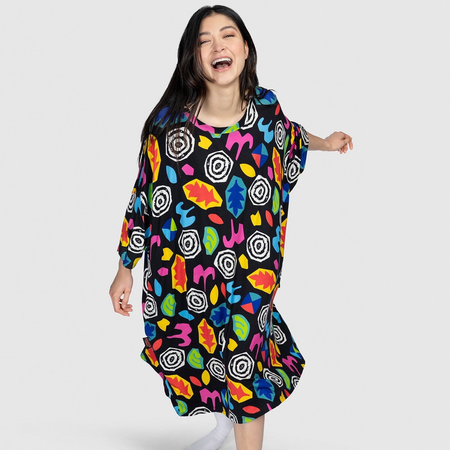 Smiling woman wearing a loose black dress with colorful abstract shapes on a plain light gray background