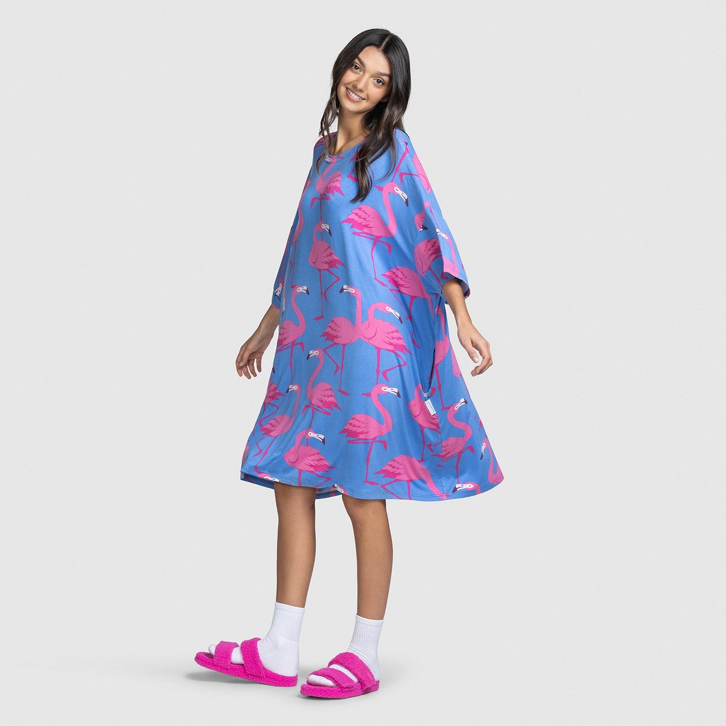 Woman wearing a blue dress with pink flamingo prints and pink slippers, smiling and posing against a plain background.