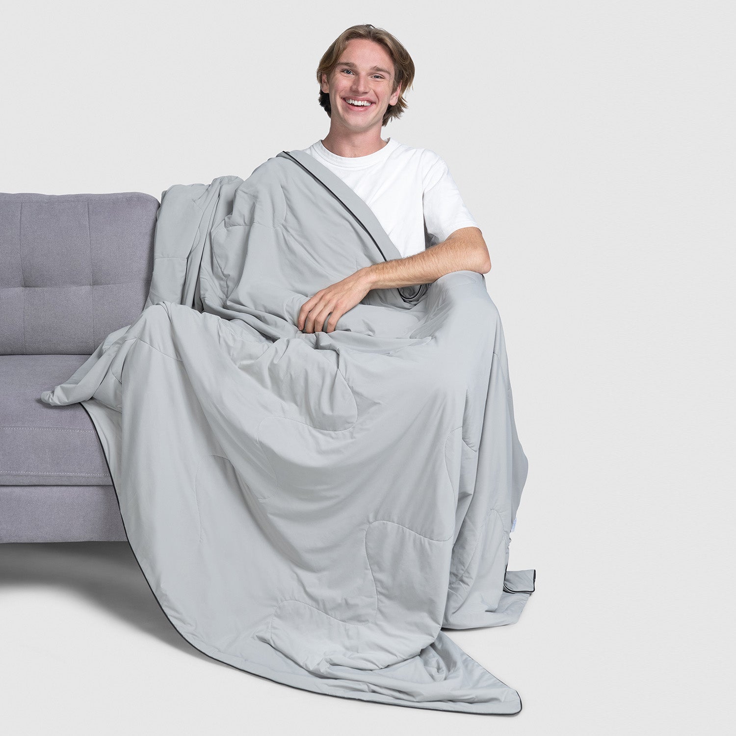Young man sitting on a gray couch wrapped in a large gray blanket, smiling at the camera.