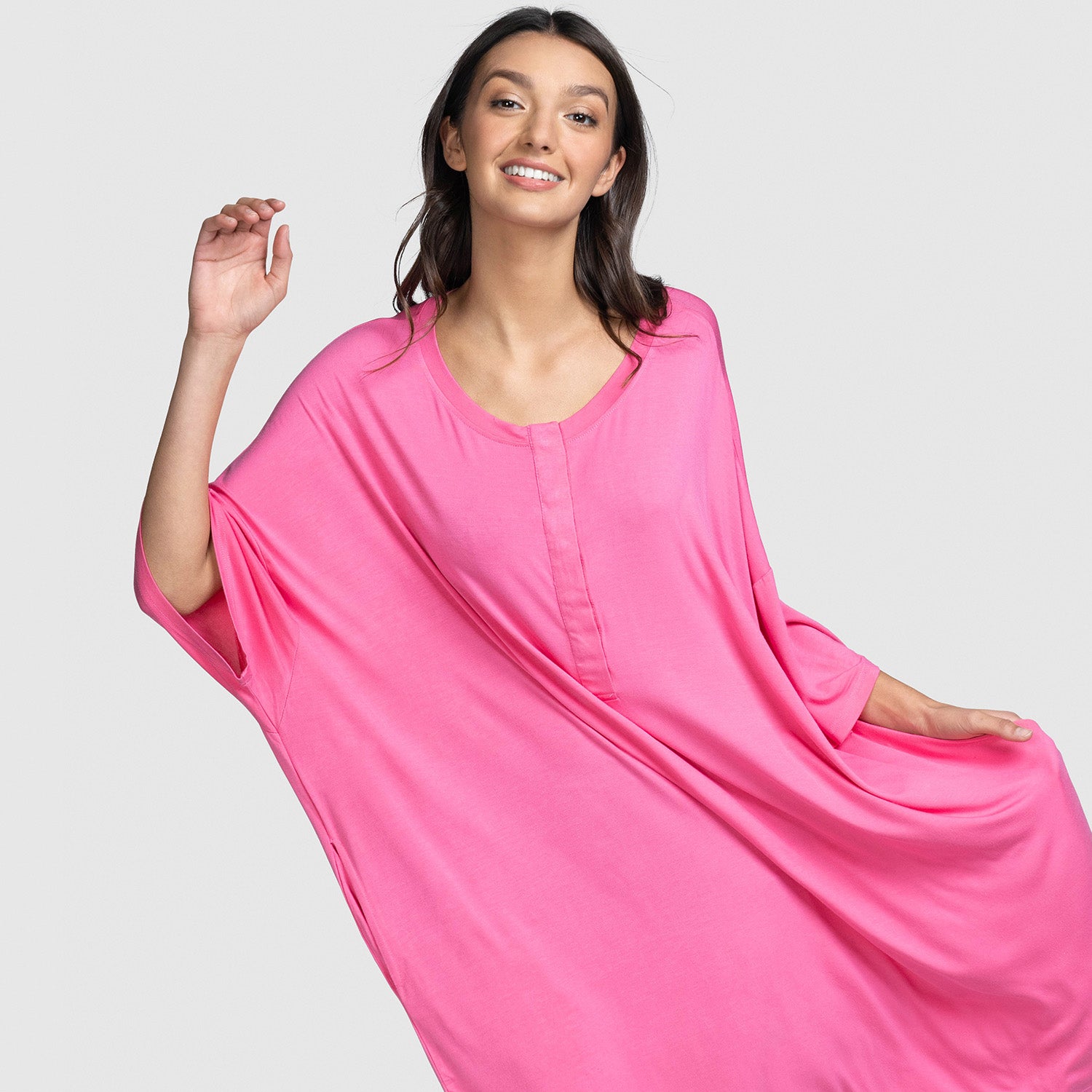 Woman smiling wearing a loose bright pink dress with wide sleeves and a round neckline, posing with one arm raised and the other holding the dress.