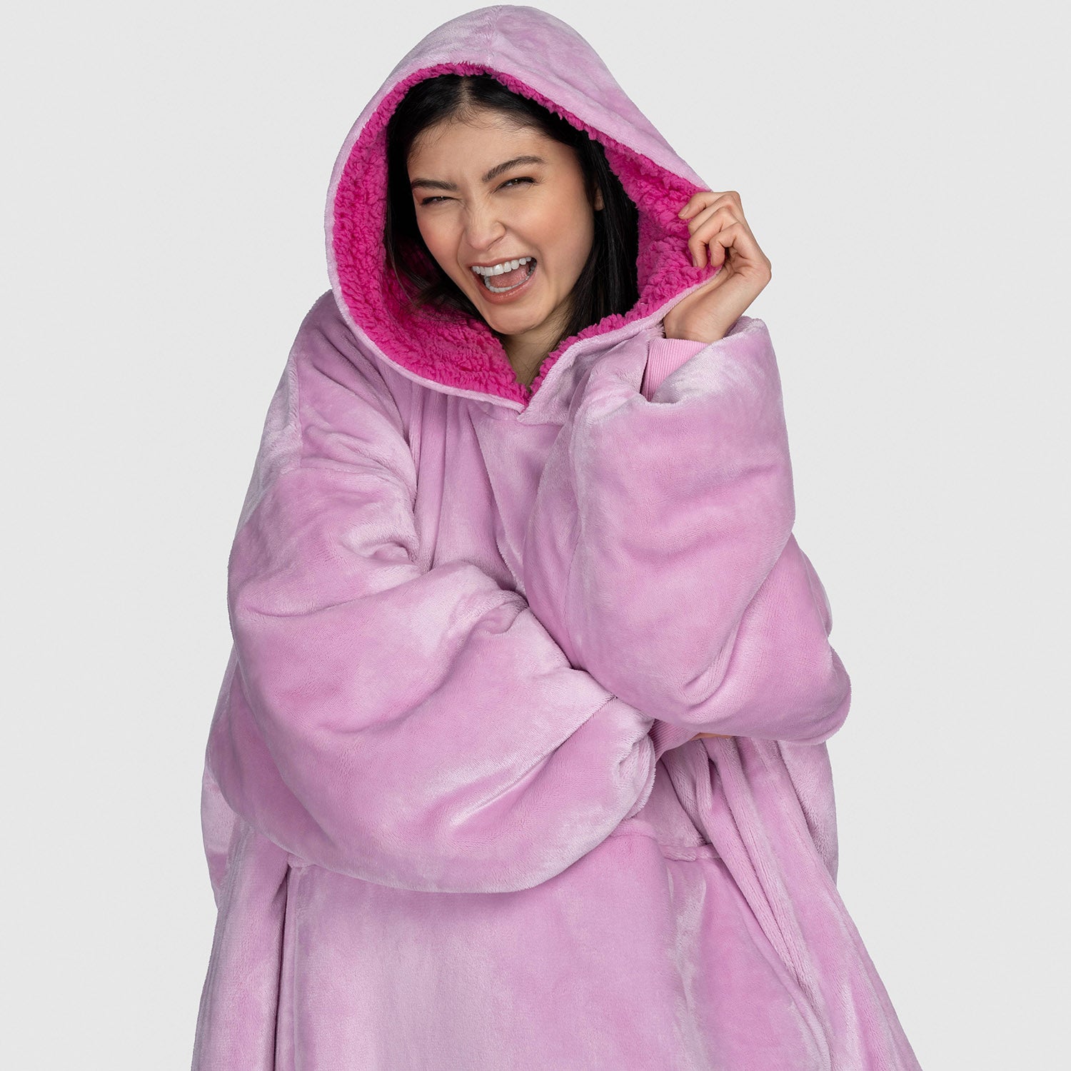 Woman wearing a large pink plush hooded garment, smiling and holding the hood; model is 5'2" and wears size OOD size: Shanti is 5'2" wearing a size OOD