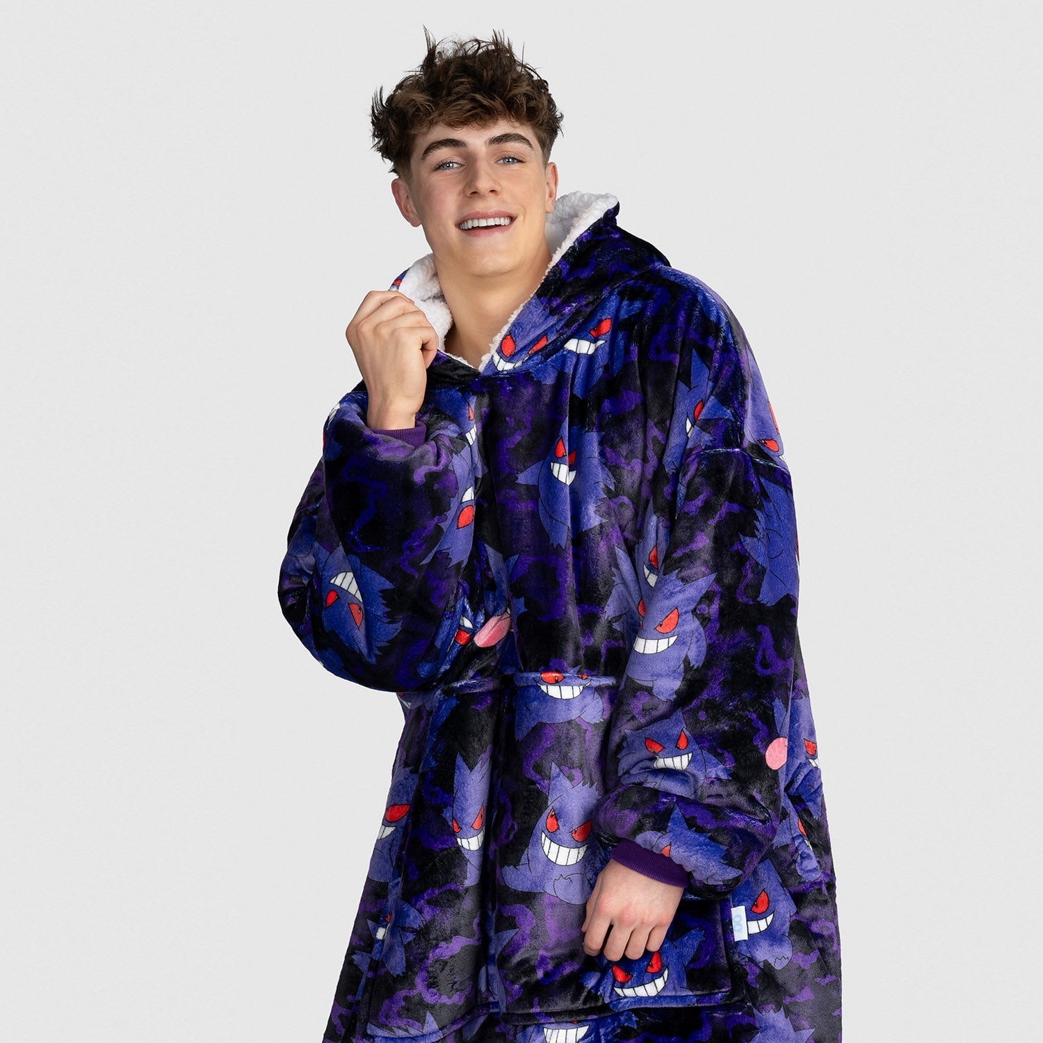 Young man wearing a hooded blanket covered in multiple purple Pokémon Gengar faces with red eyes and wide grins.