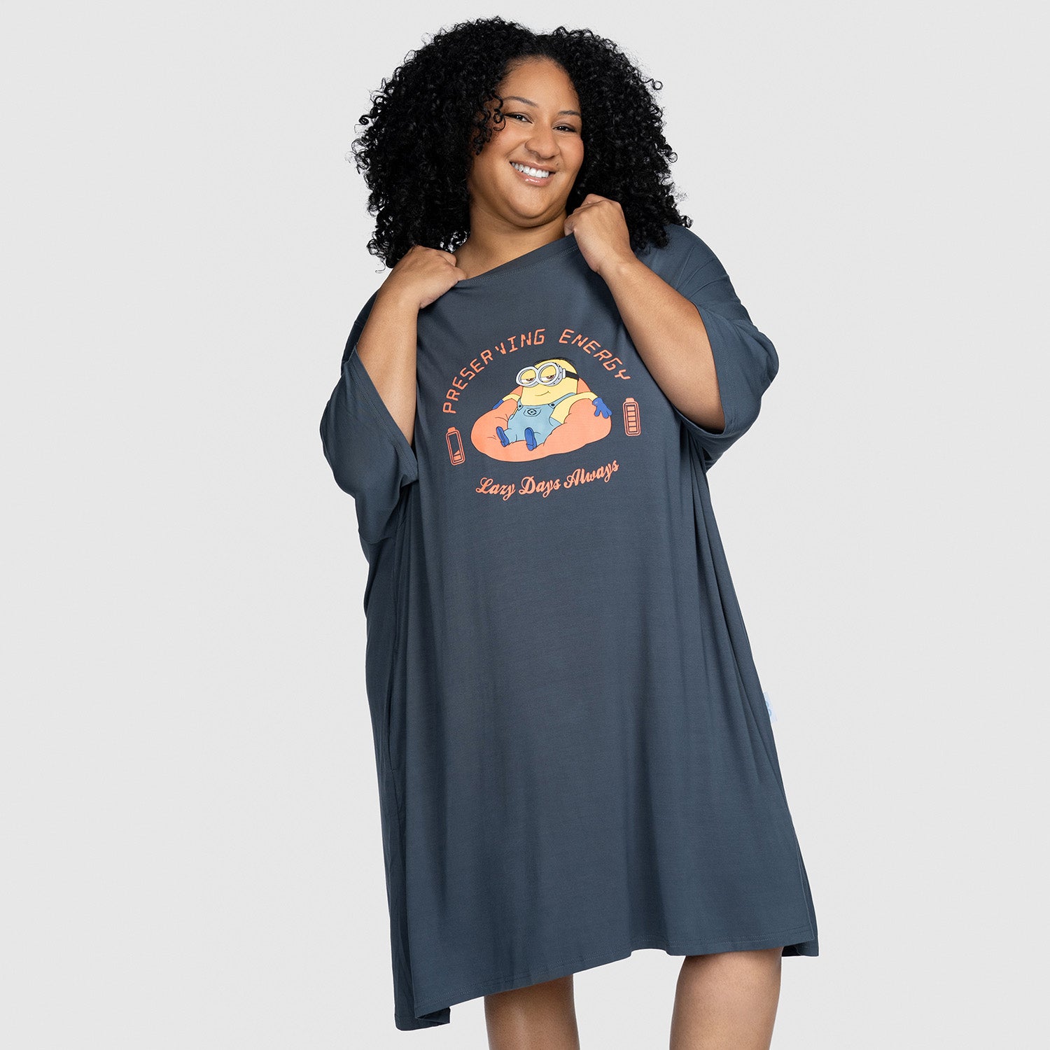 Smiling woman wearing a loose dark gray dress with a Minion character and the text 'Preserving Energy Lazy Days Always' on the front