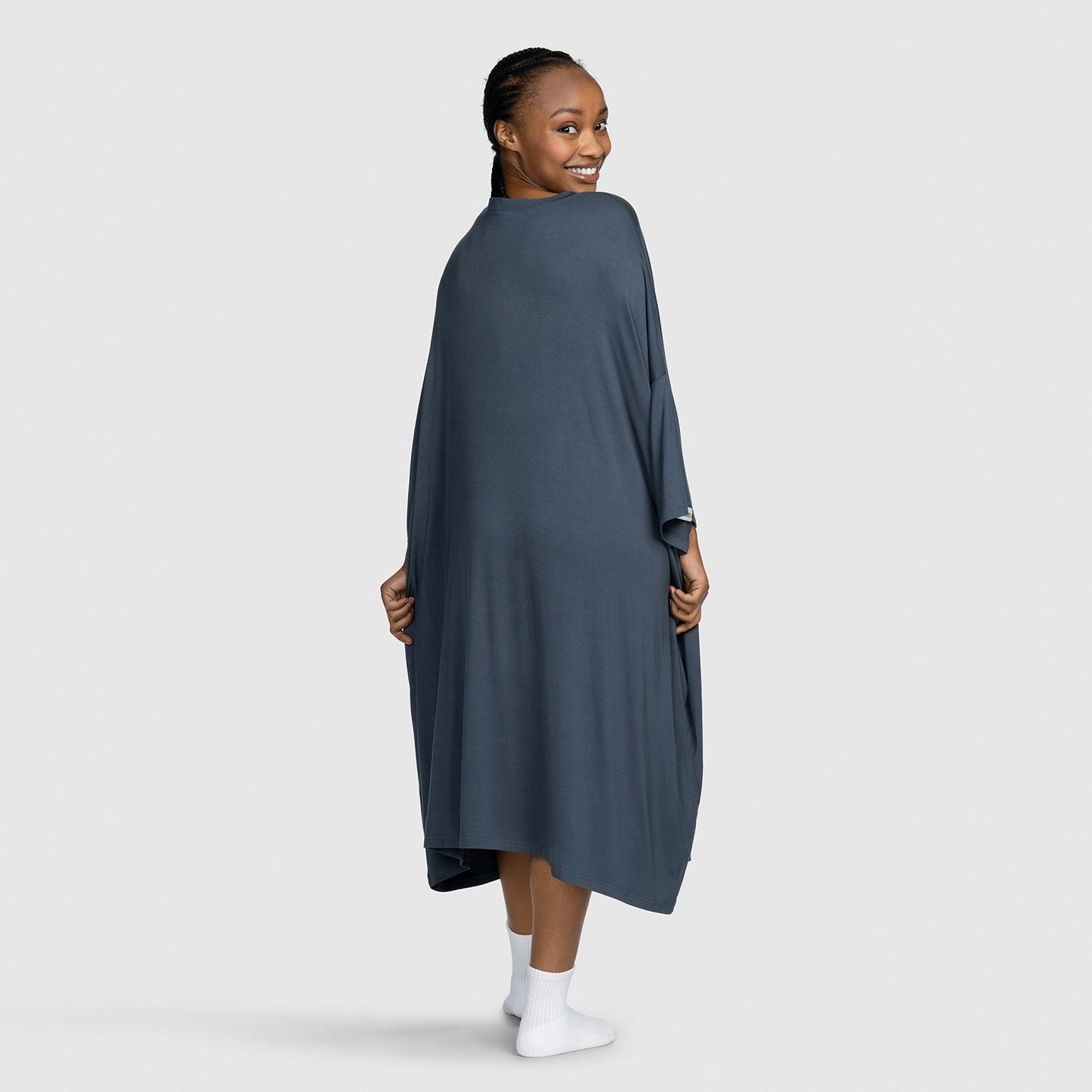 Smiling woman wearing a dark gray shawl or wrap, viewed from behind, standing on a light gray background.