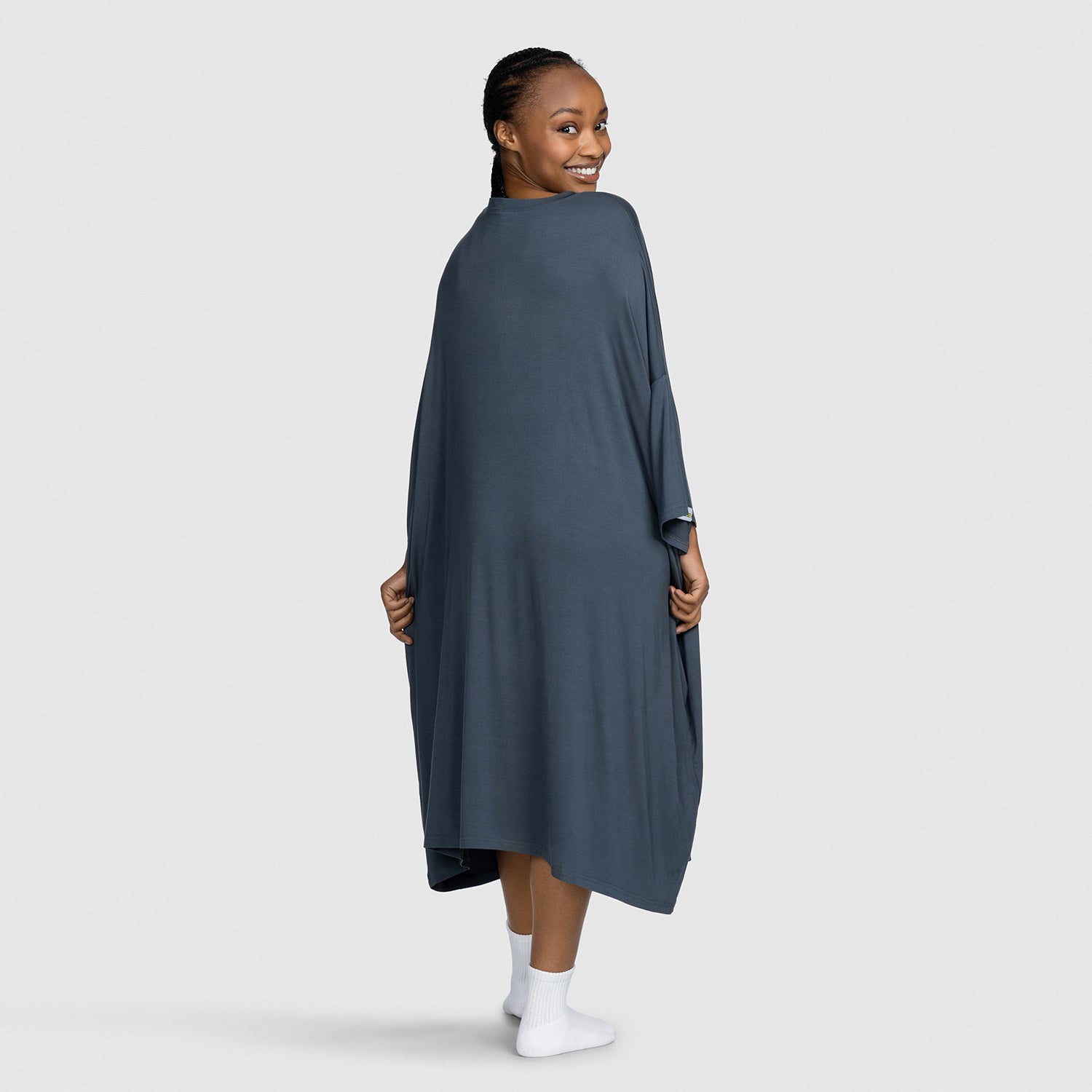 Smiling woman wearing a dark gray shawl or wrap, viewed from behind, standing on a light gray background.
