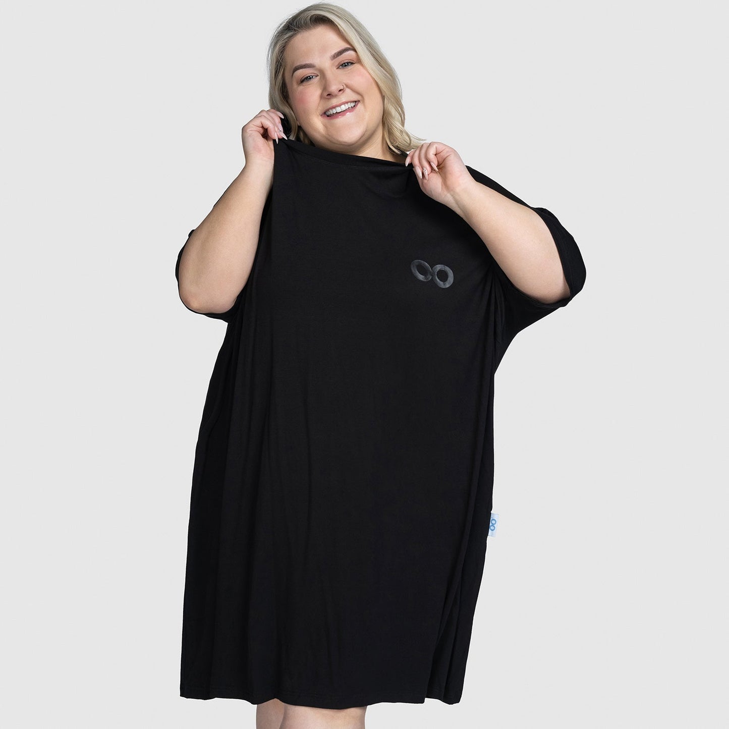Smiling plus-size woman holding a loose black dress with a small circular logo on the chest