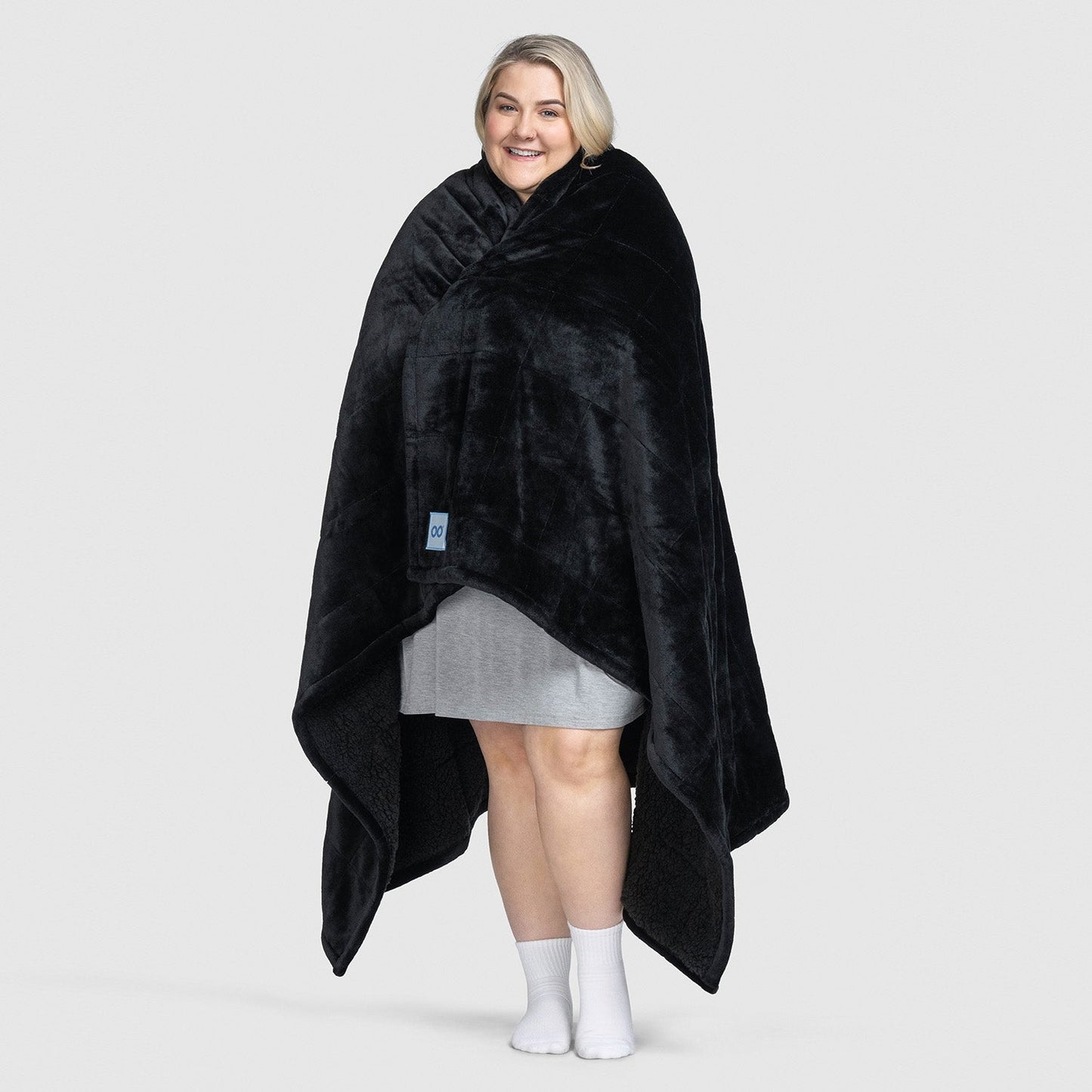 Person wrapped in a black plush blanket, standing and smiling, wearing a gray dress and white socks.