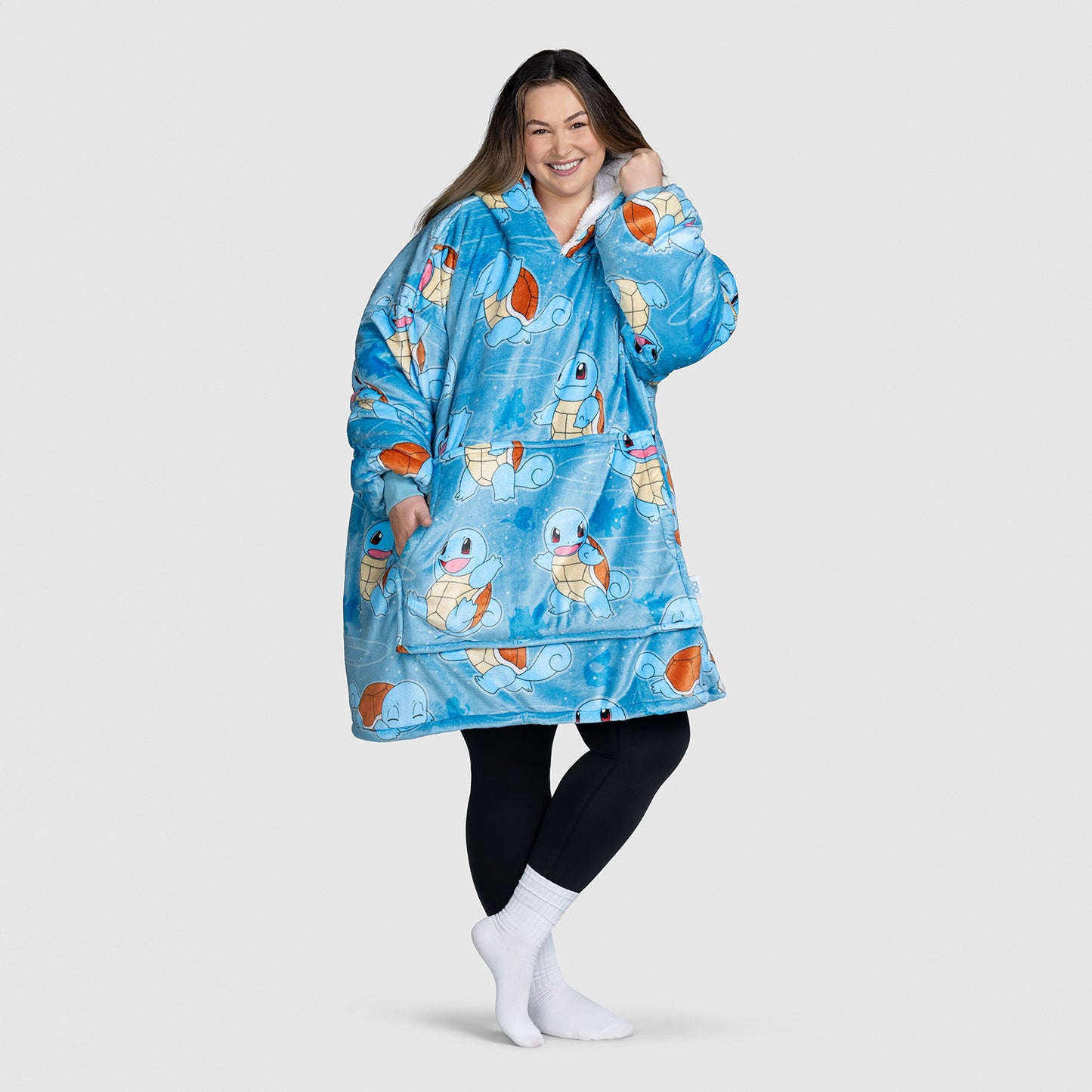 Woman wearing a blue wearable blanket with Pokémon Squirtle patterns, smiling and posing with one hand near her face.