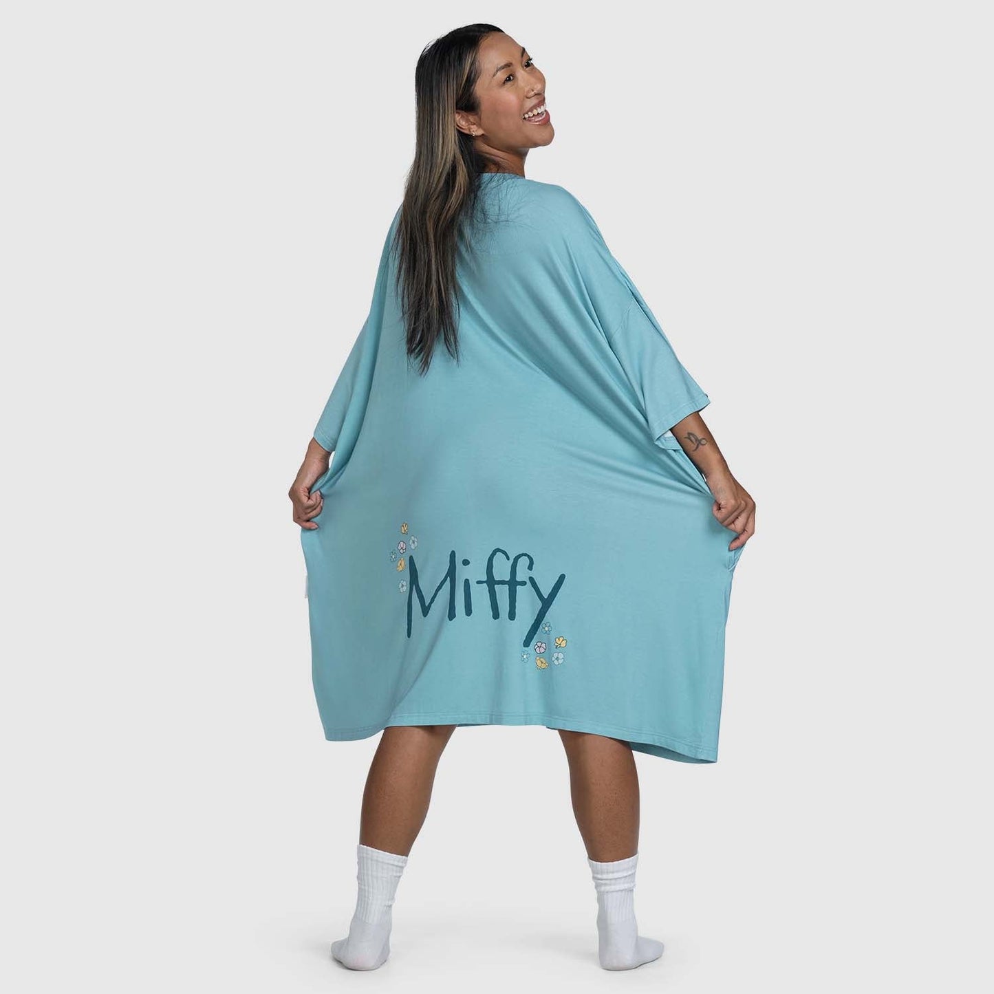 Woman wearing a blue garment with the word 'Miffy' and small flower designs on the back, smiling and looking over her shoulder.