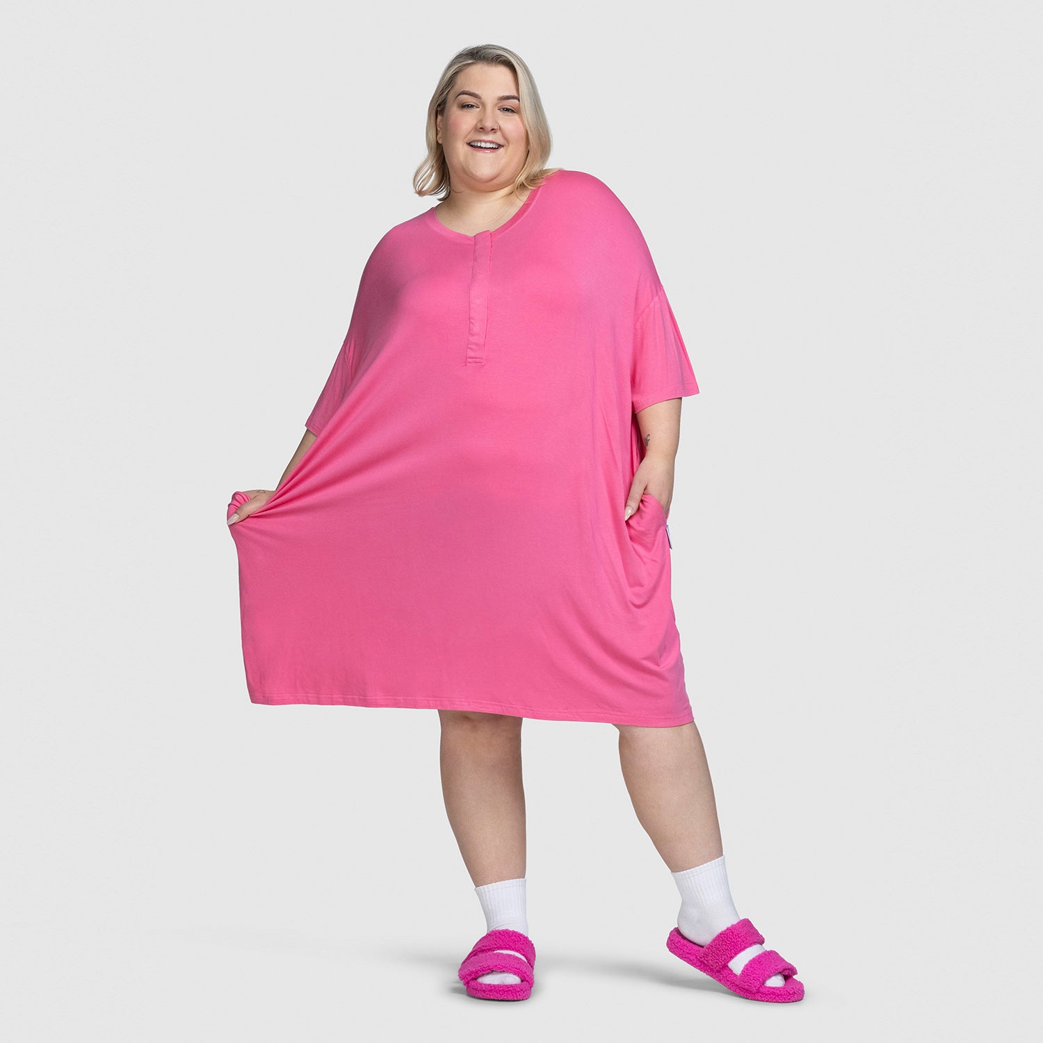 Smiling plus-size woman wearing a bright pink loose dress and matching pink fuzzy slippers, standing against a plain background.