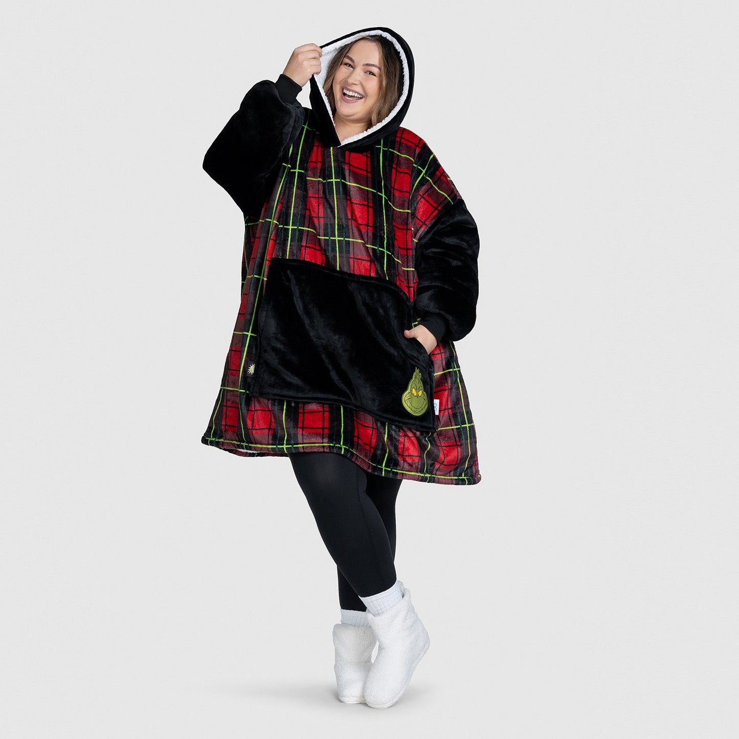 Smiling woman wearing a red and black plaid oversized hoodie with a black front pocket and a Grinch patch, posing with one hand holding the hood.