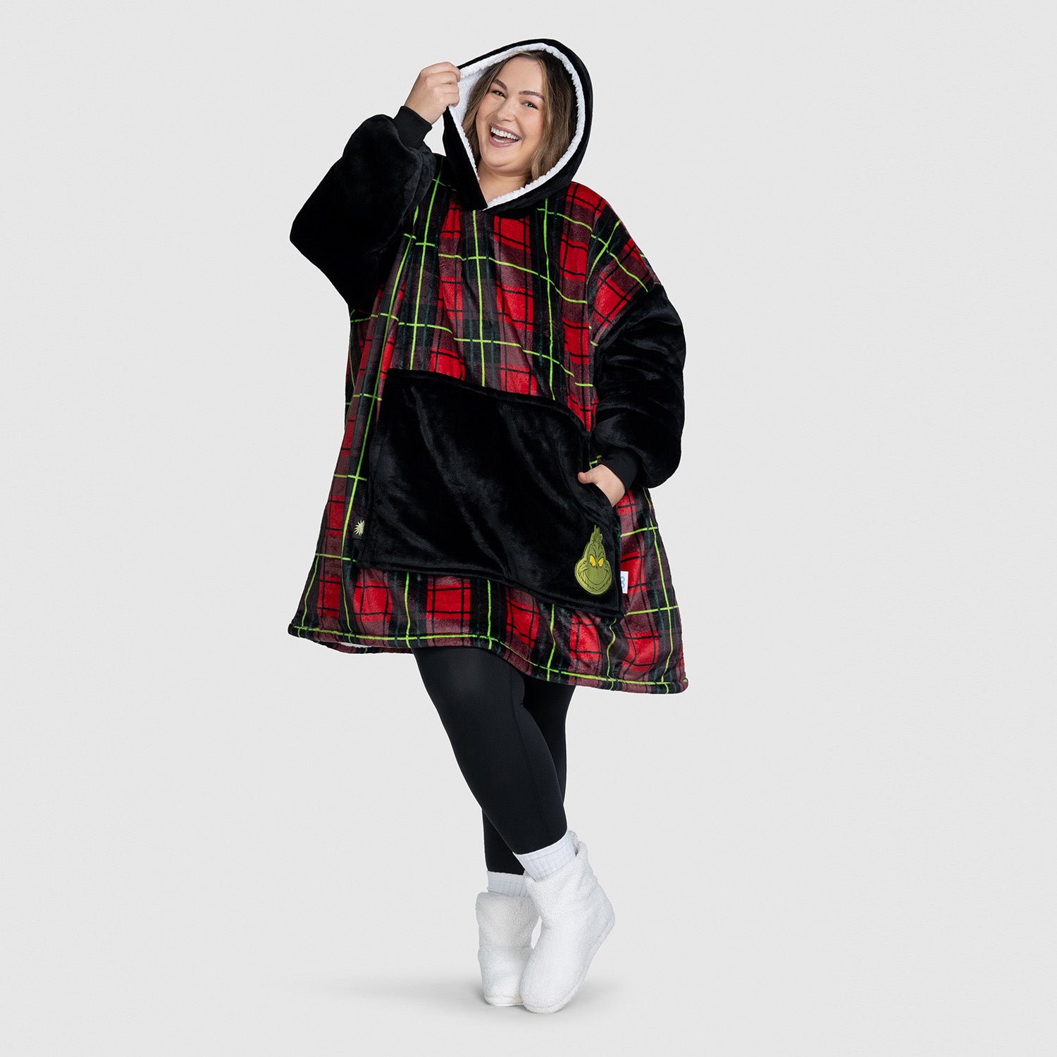 Smiling woman wearing a red and black plaid oversized hoodie with a black front pocket and a Grinch patch, posing with one hand holding the hood.