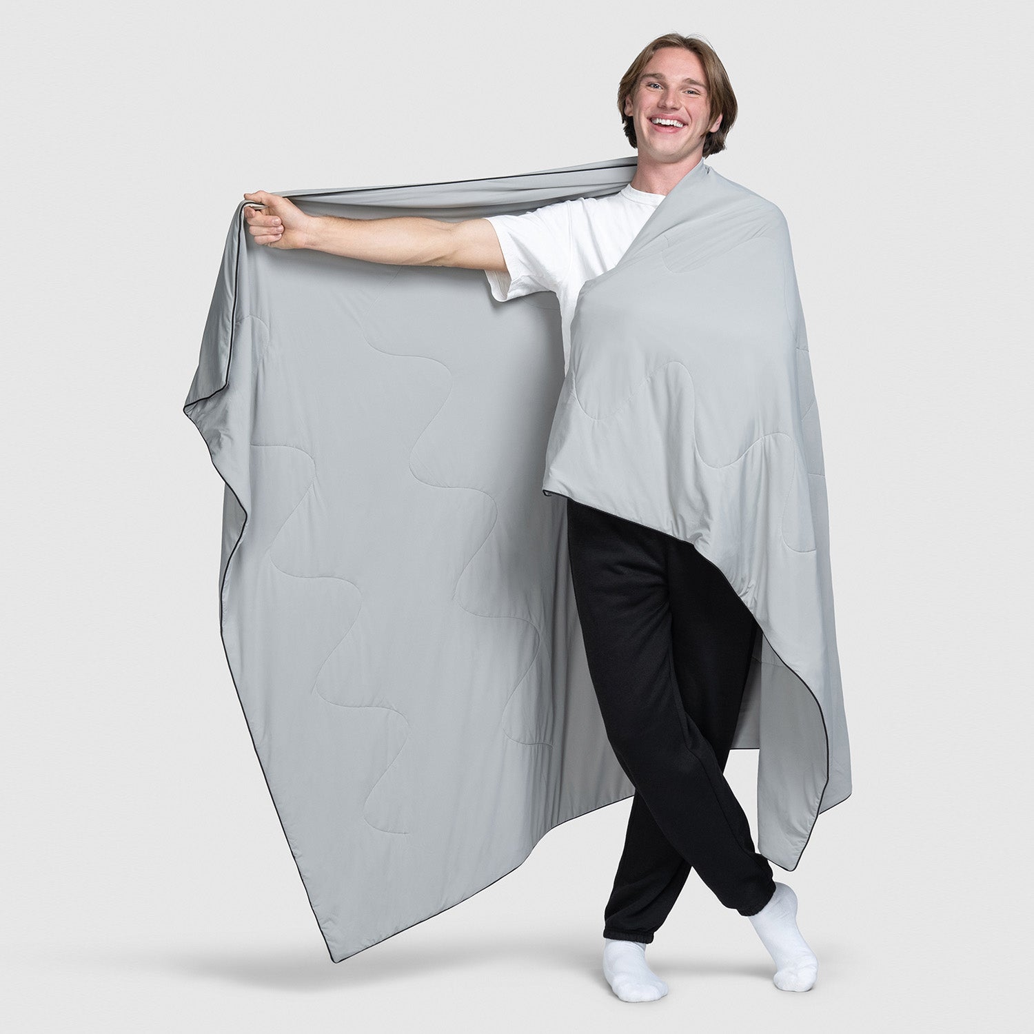 Smiling person standing wrapped in a large grey blanket or fabric, holding it out with one arm extended.