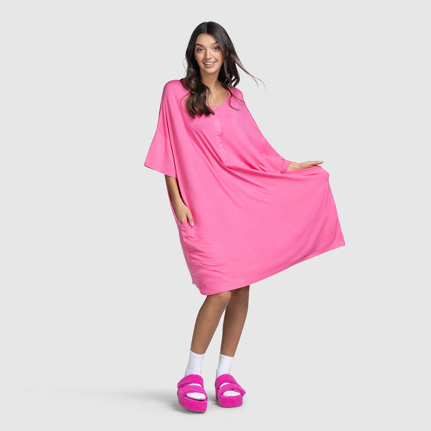 Woman wearing a bright pink loose dress and matching slippers, smiling and holding the dress out to the side.