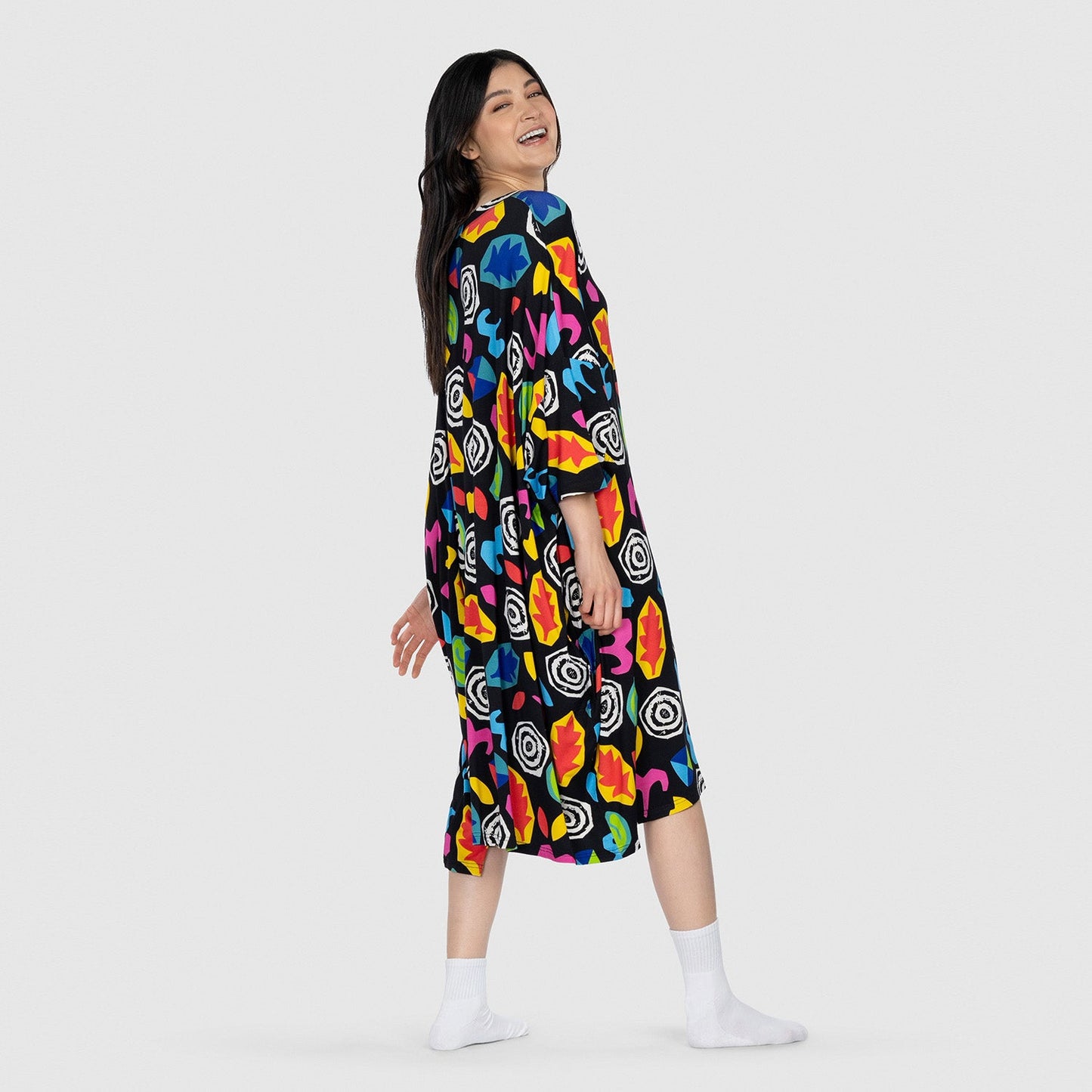 Woman smiling and looking back, wearing a colorful abstract patterned dress and white socks, standing against a plain light gray background.