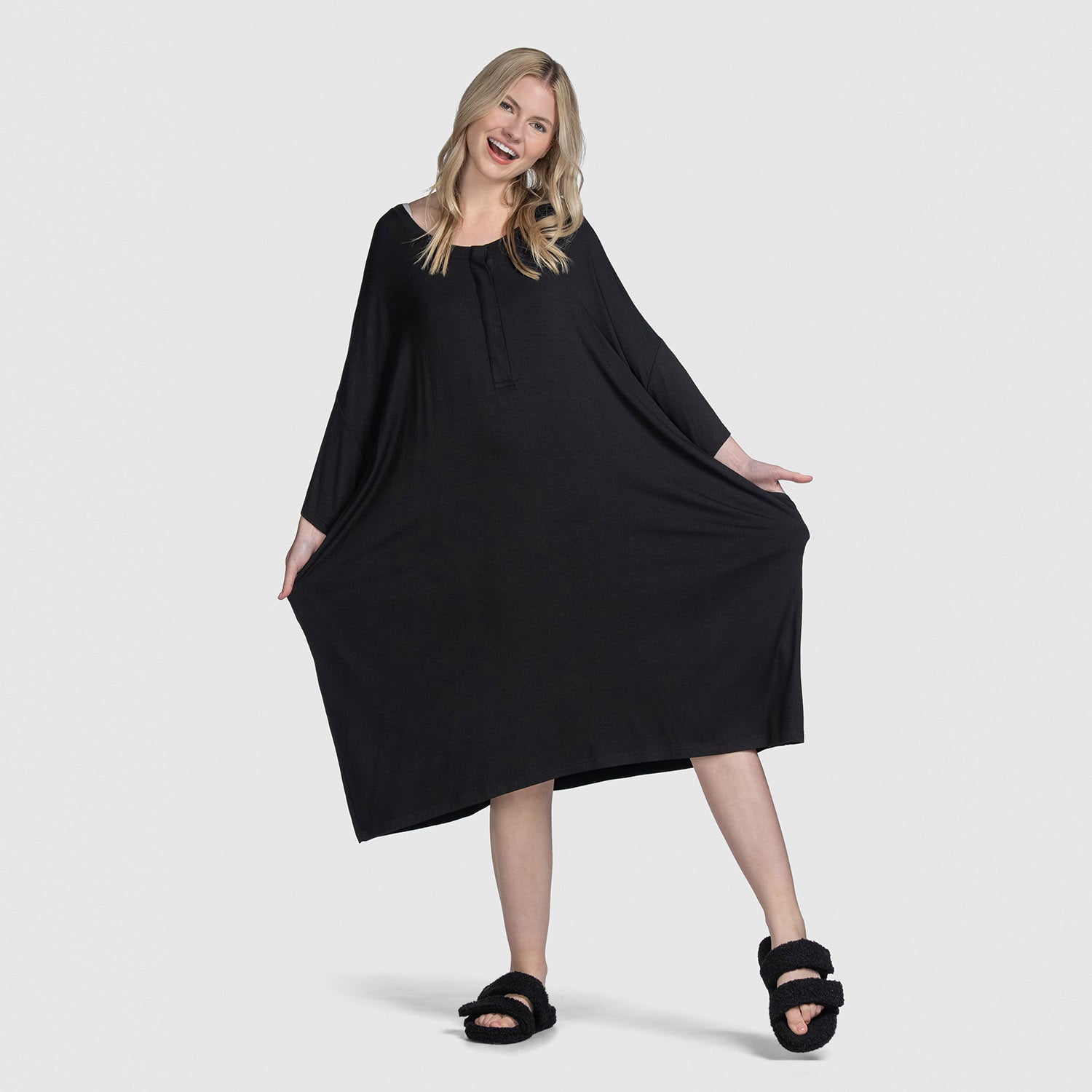 Woman smiling and posing wearing a loose black long-sleeve dress and black slippers against a plain light gray background.