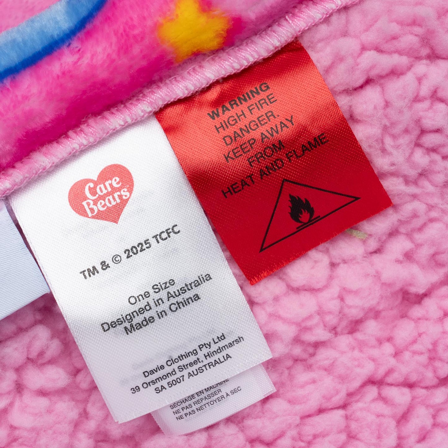 Clothing tags on pink fabric showing Care Bears logo, trademark info, manufacturing details, and a red warning label with fire hazard symbol and text: WARNING HIGH FIRE DANGER KEEP AWAY FROM HEAT AND FLAME