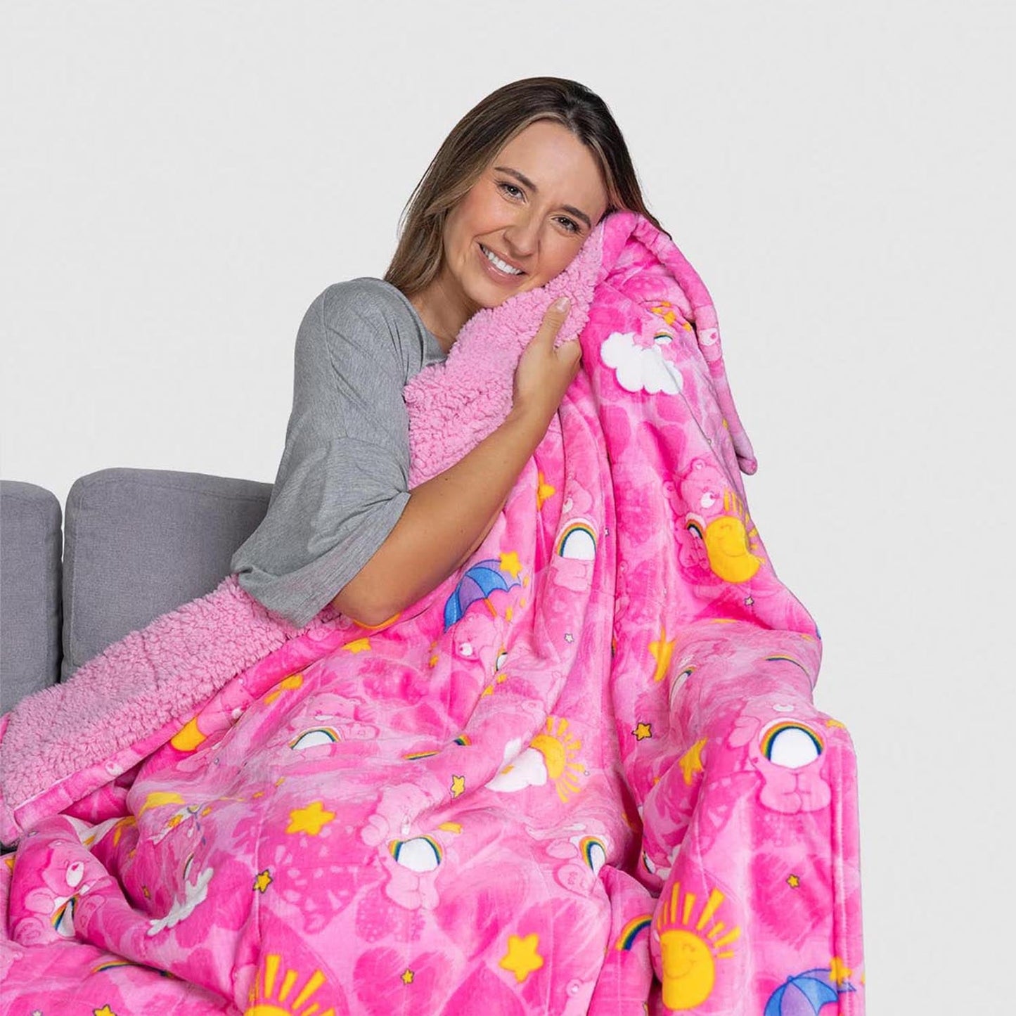 A smiling woman hugging a pink Care Bears patterned blanket while sitting on a gray couch.