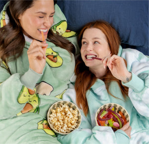 Two women wearing avocado-patterned loungewear, lying on a bed, smiling and eating popcorn and gummy candies.