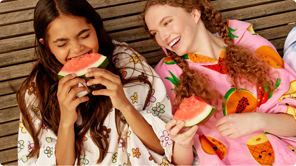 Two women wearing colorful fruit-patterned hooded blankets eating watermelon slices and smiling on wooden floor.