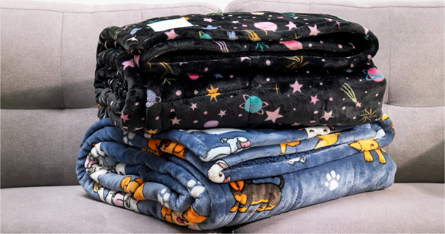 Two folded fleece blankets with colorful patterns, one with space motifs and the other with dog and paw prints, on a gray couch