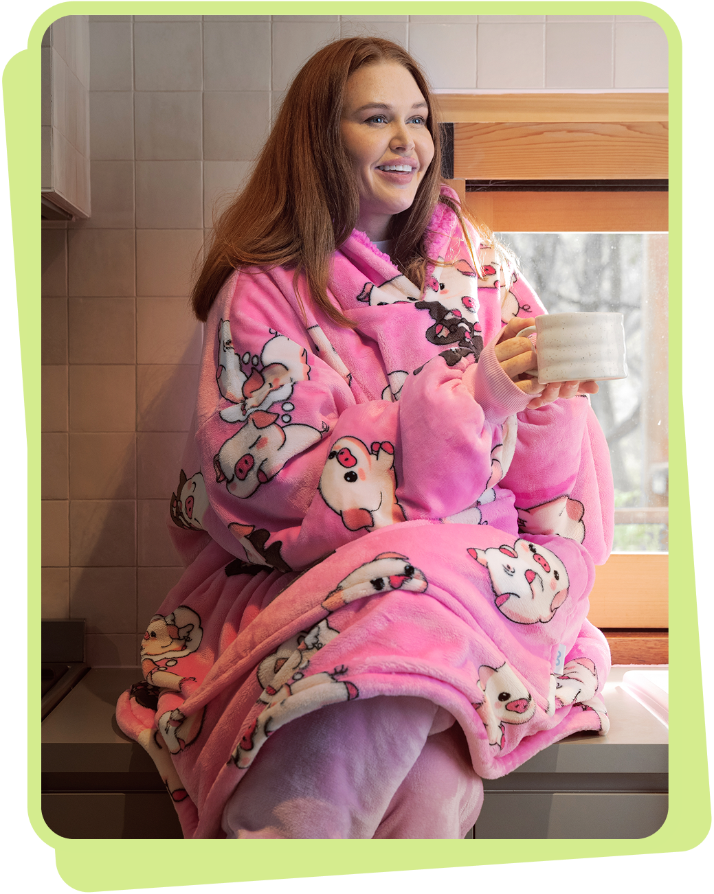 Woman wearing a pink pig-patterned Oodie robe holding a white mug, sitting by a window.