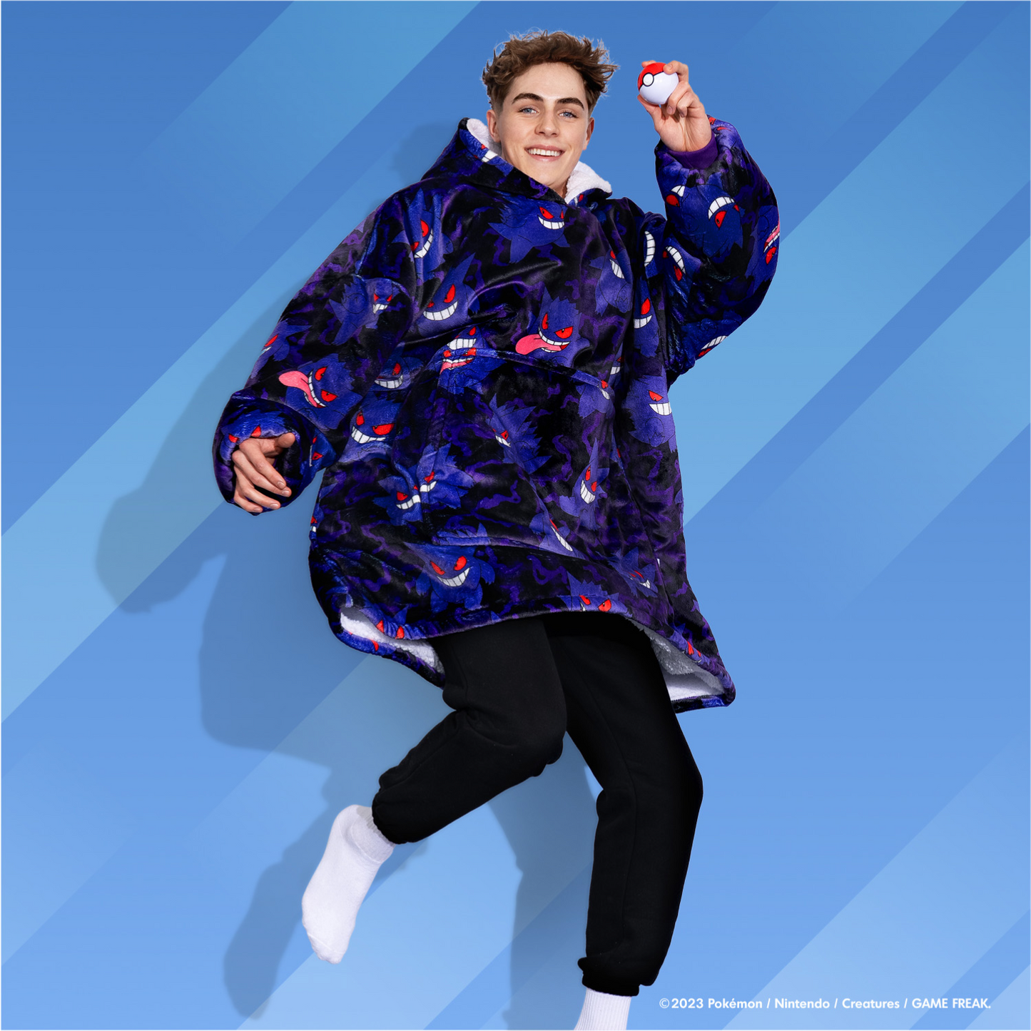 Person jumping wearing a purple oversized hoodie covered with Gengar faces and holding a Poké Ball against a blue background