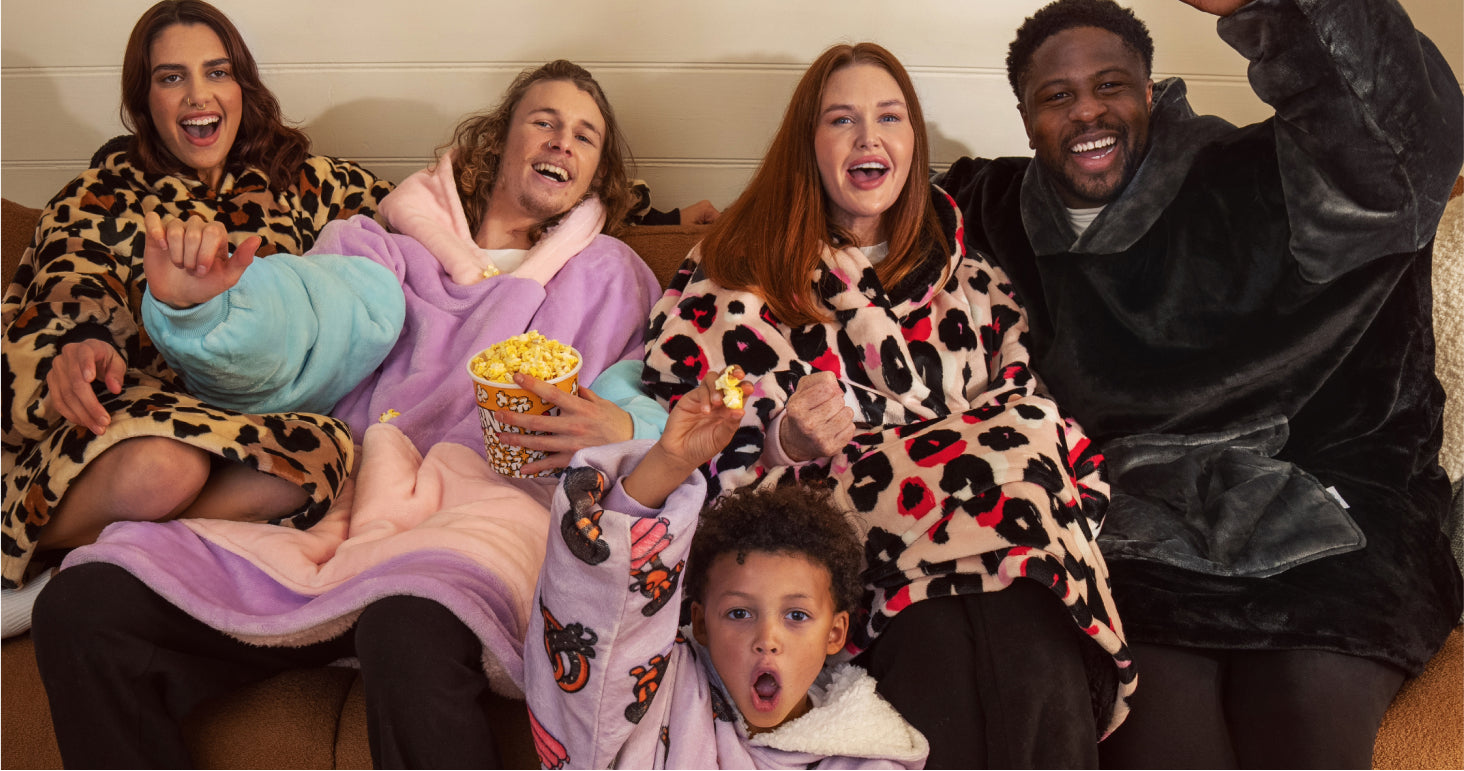 Five people wearing colorful fleece hoodies sitting on a couch, smiling and holding popcorn, enjoying a cozy moment.