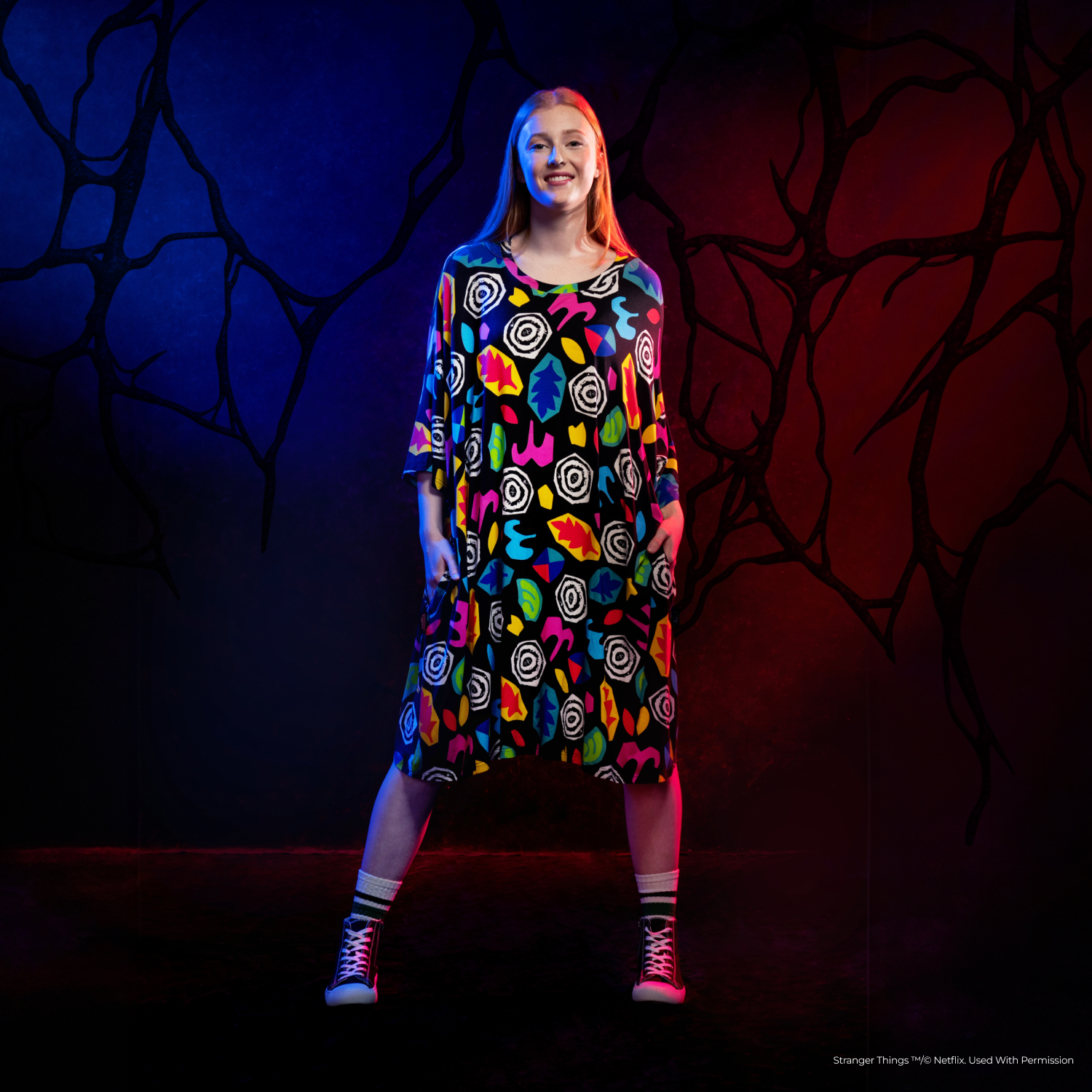 Young woman standing confidently in a colorful patterned dress against a dark background with abstract black shapes and red and blue lighting.