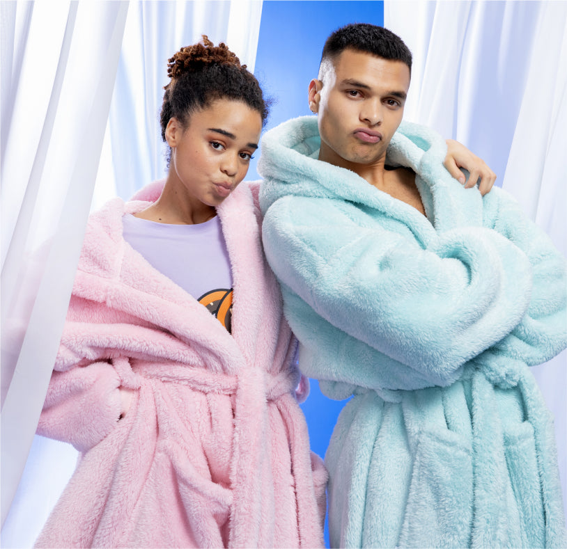 Two people posing in fluffy robes, one in pink and one in light blue, against a blue and white curtain background.