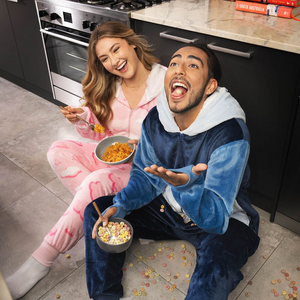 Two people sitting on a kitchen floor in cozy loungewear eating cereal, one spilling some on the floor, smiling and looking up.