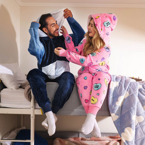 Two people sitting on a bed wearing cozy onesies, one in navy and white, the other in pink with colorful heart patterns.
