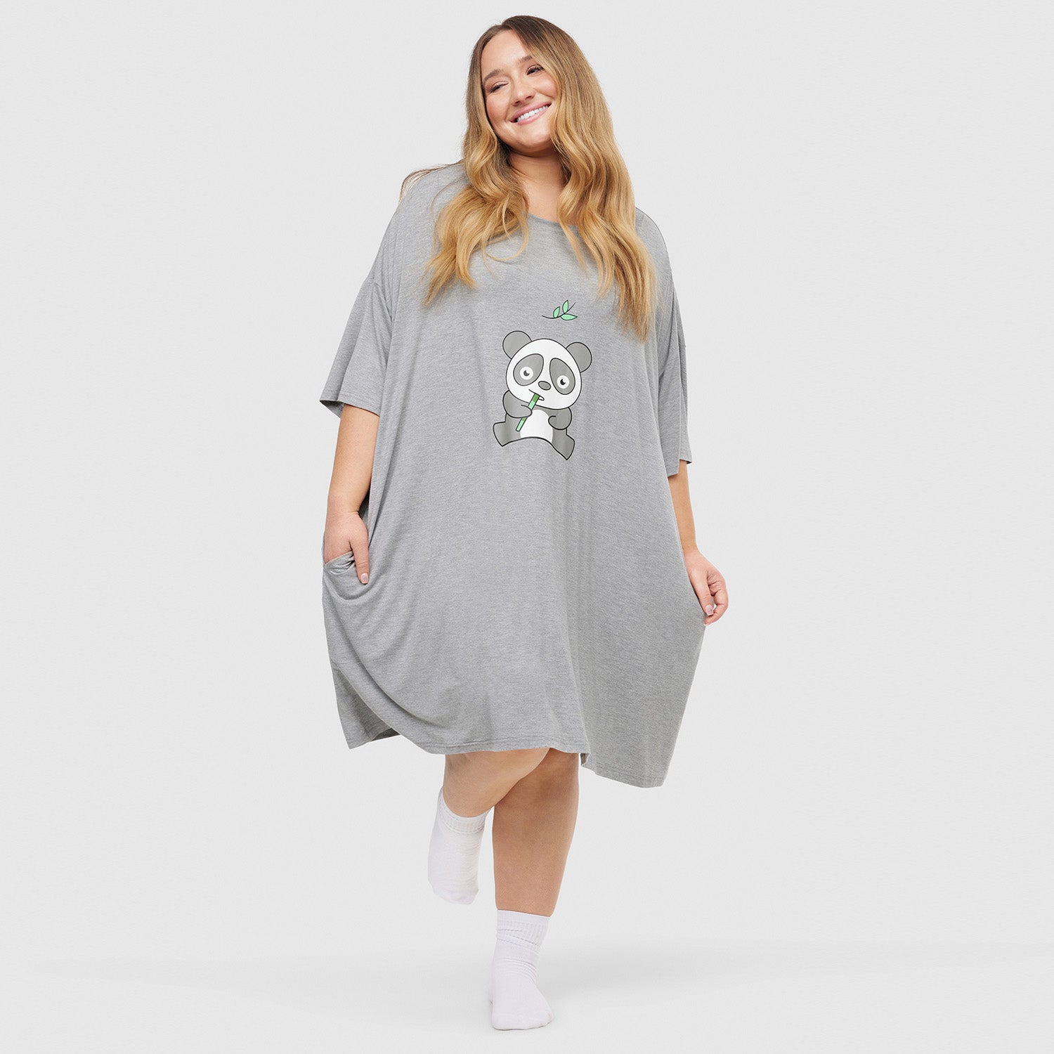 Woman wearing a loose gray dress with a cartoon panda holding bamboo printed on the front, smiling and posing with one foot raised, wearing white socks.