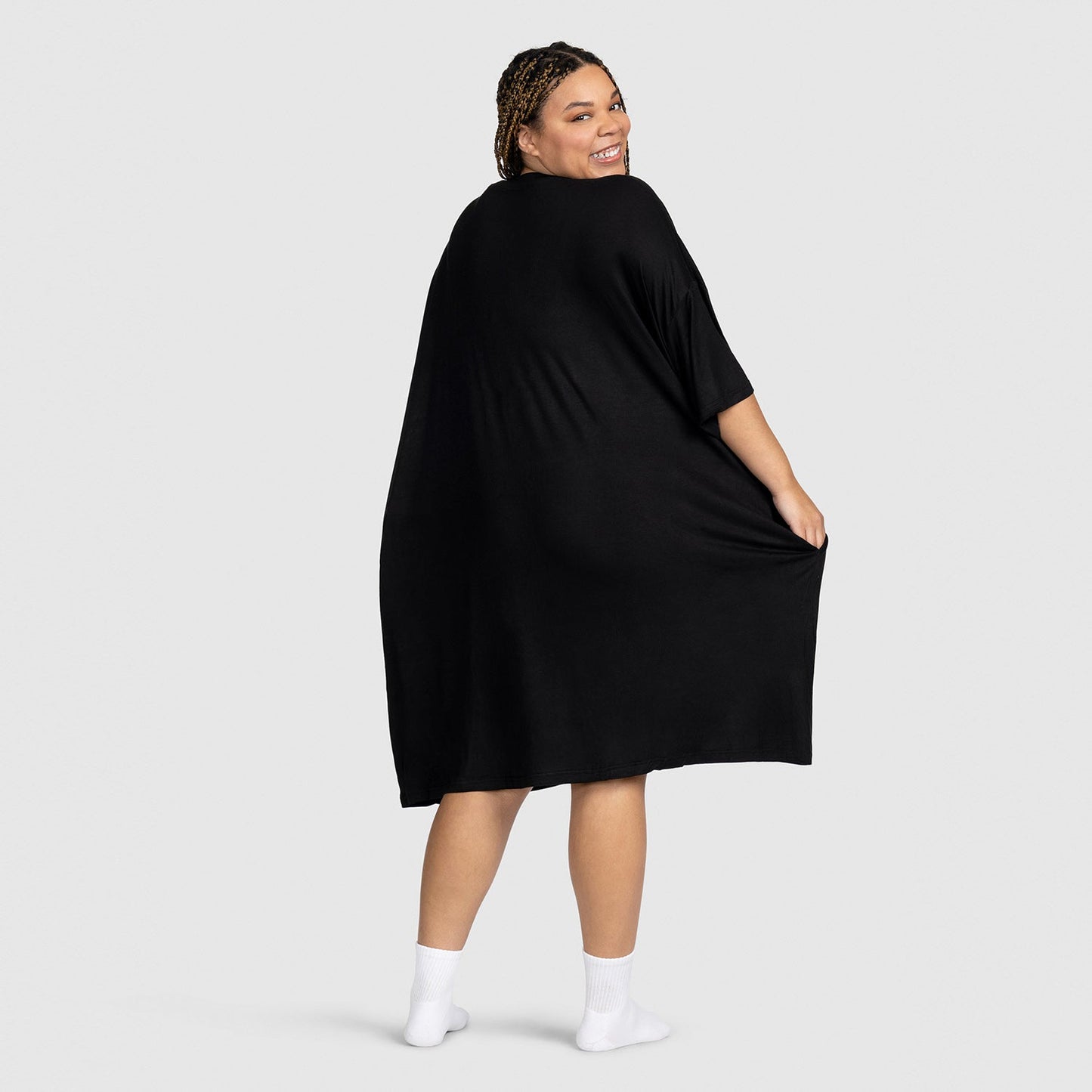 Smiling woman wearing a loose black dress, standing barefoot on a light gray background, facing away and looking over her shoulder.