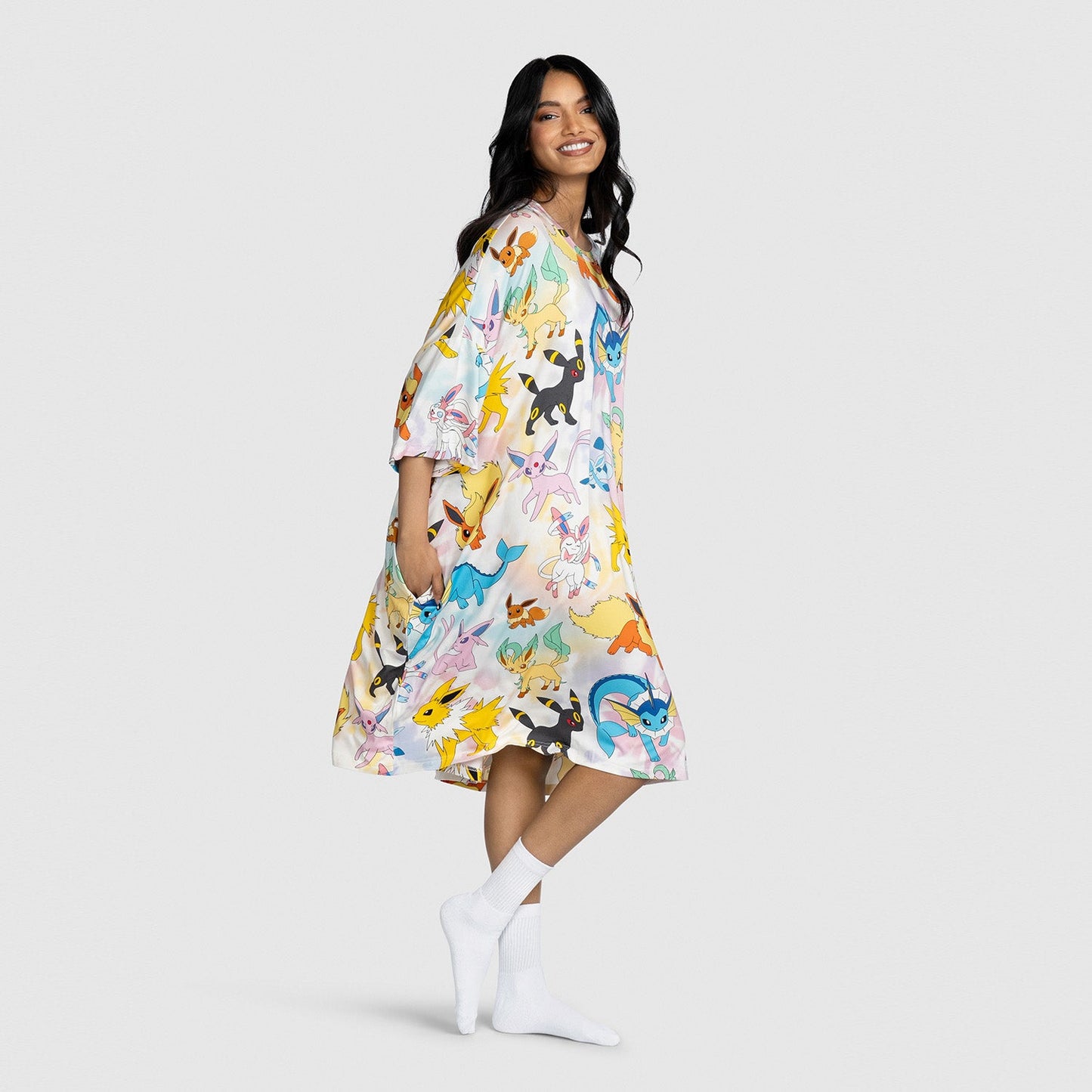 Woman wearing a loose, knee-length dress printed with colorful Pokémon Eevee evolution characters, standing barefoot with white socks against a plain background.