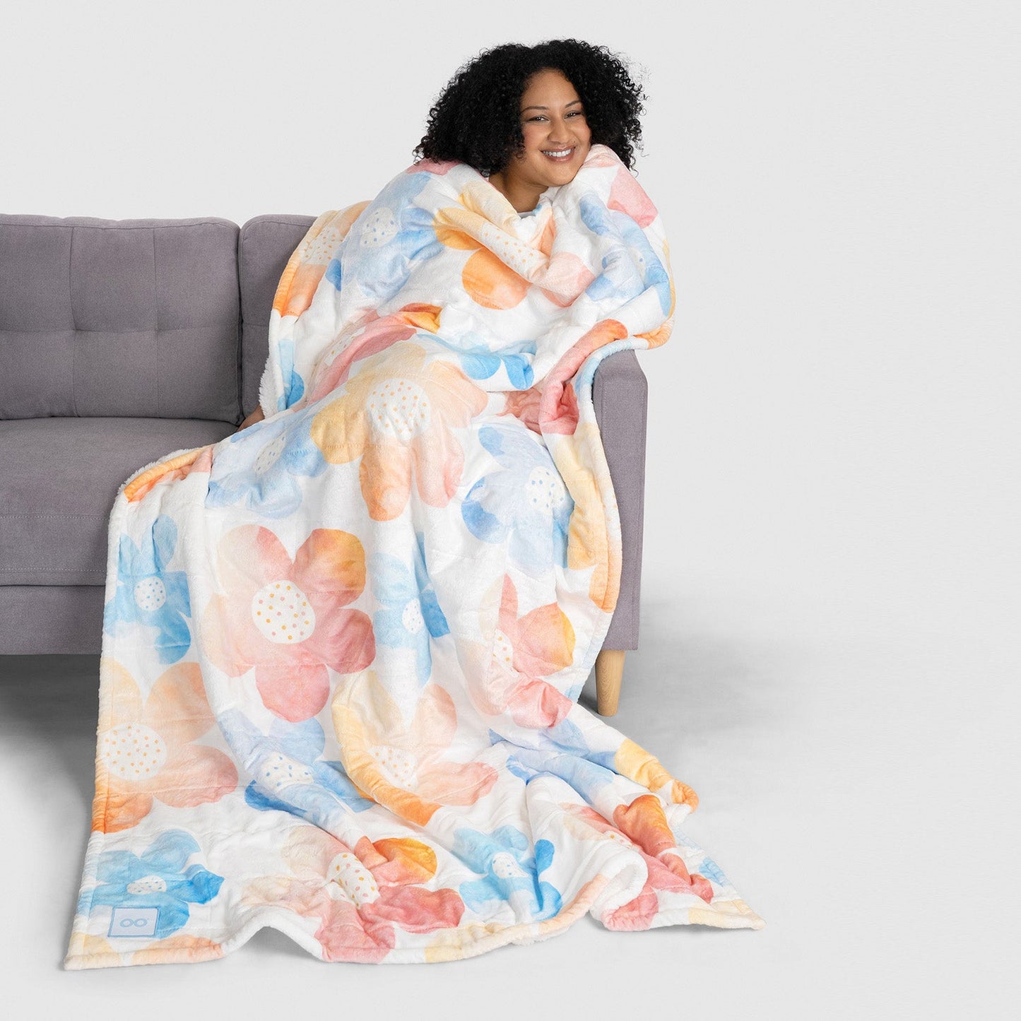Smiling woman wrapped in a large floral-patterned blanket sitting on a gray couch