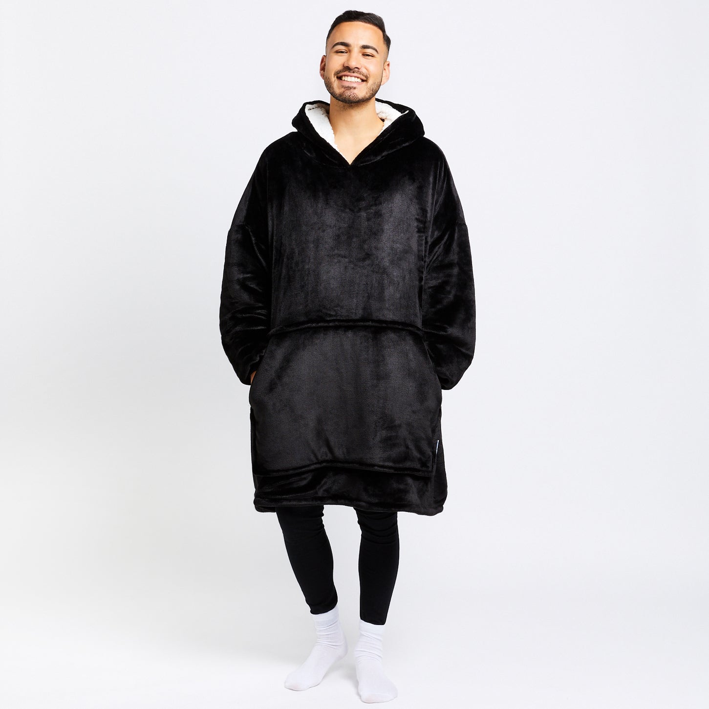 Smiling man wearing a black oversized hooded wearable blanket with pockets, standing against a white background.