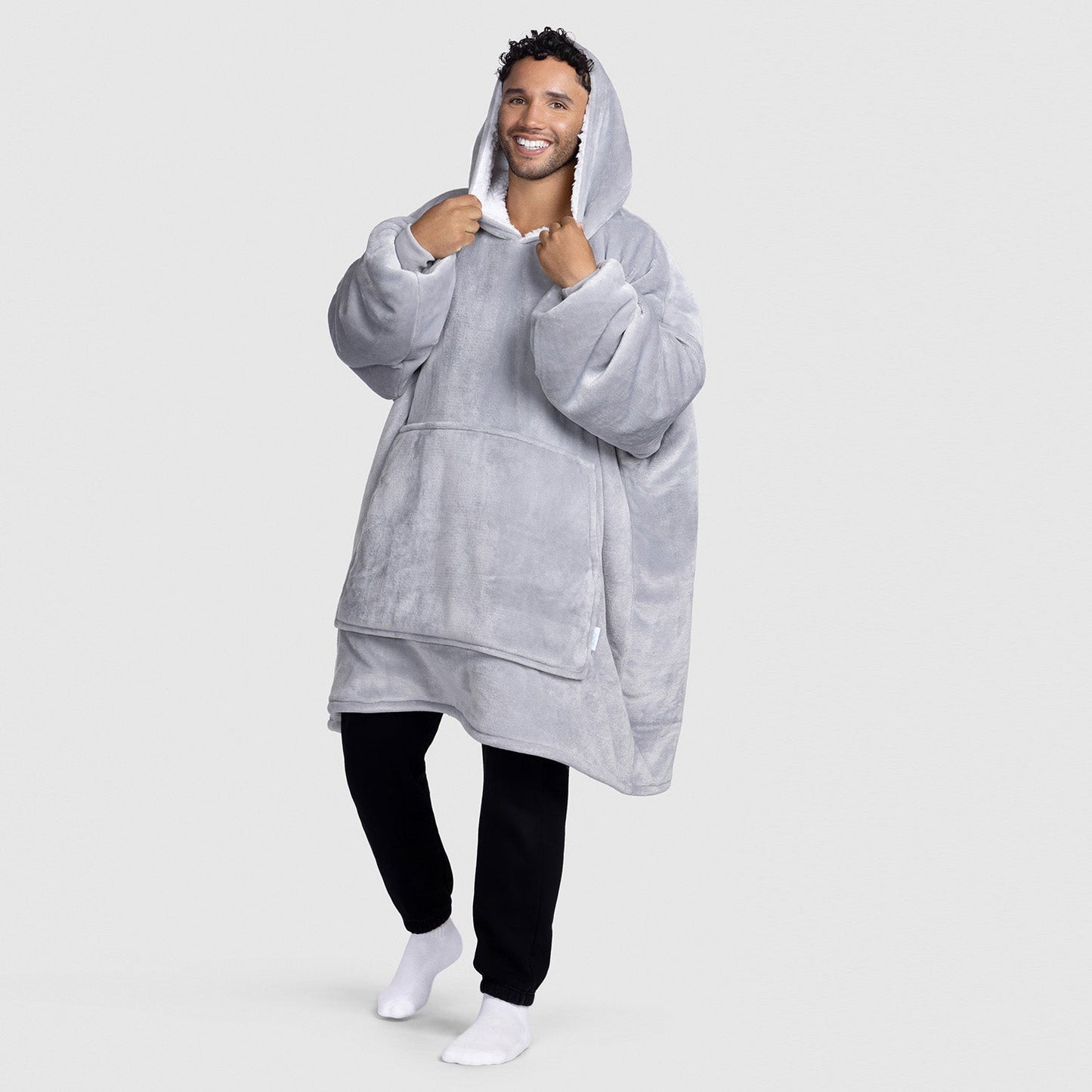 Man wearing a light gray oversized hooded wearable blanket with a front pocket, smiling and holding the hood.
