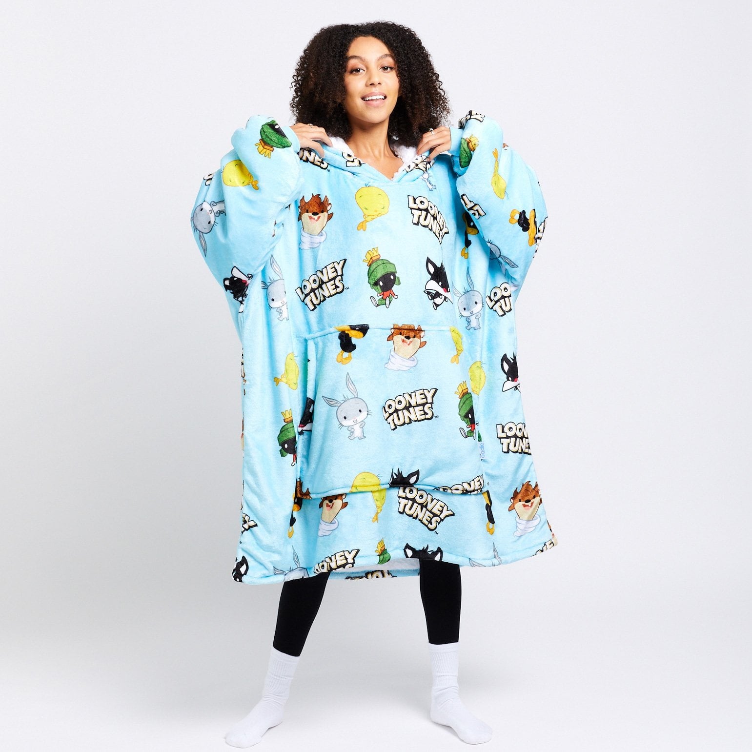 Woman wearing a blue oversized wearable blanket featuring Looney Tunes characters and logos including Bugs Bunny, Marvin the Martian, and Tweety Bird