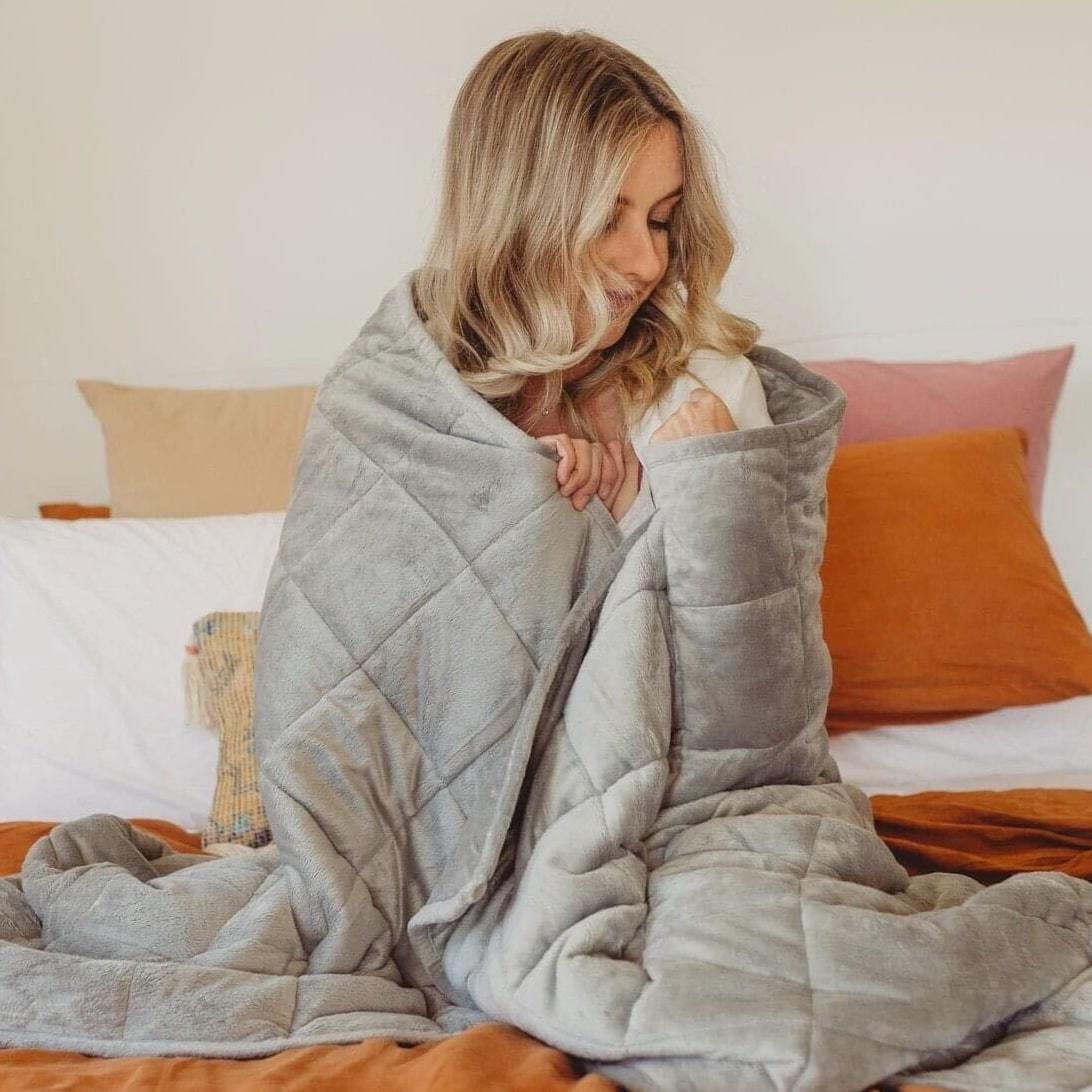 Woman wrapped in a gray quilted blanket sitting on a bed with orange and beige pillows in the background