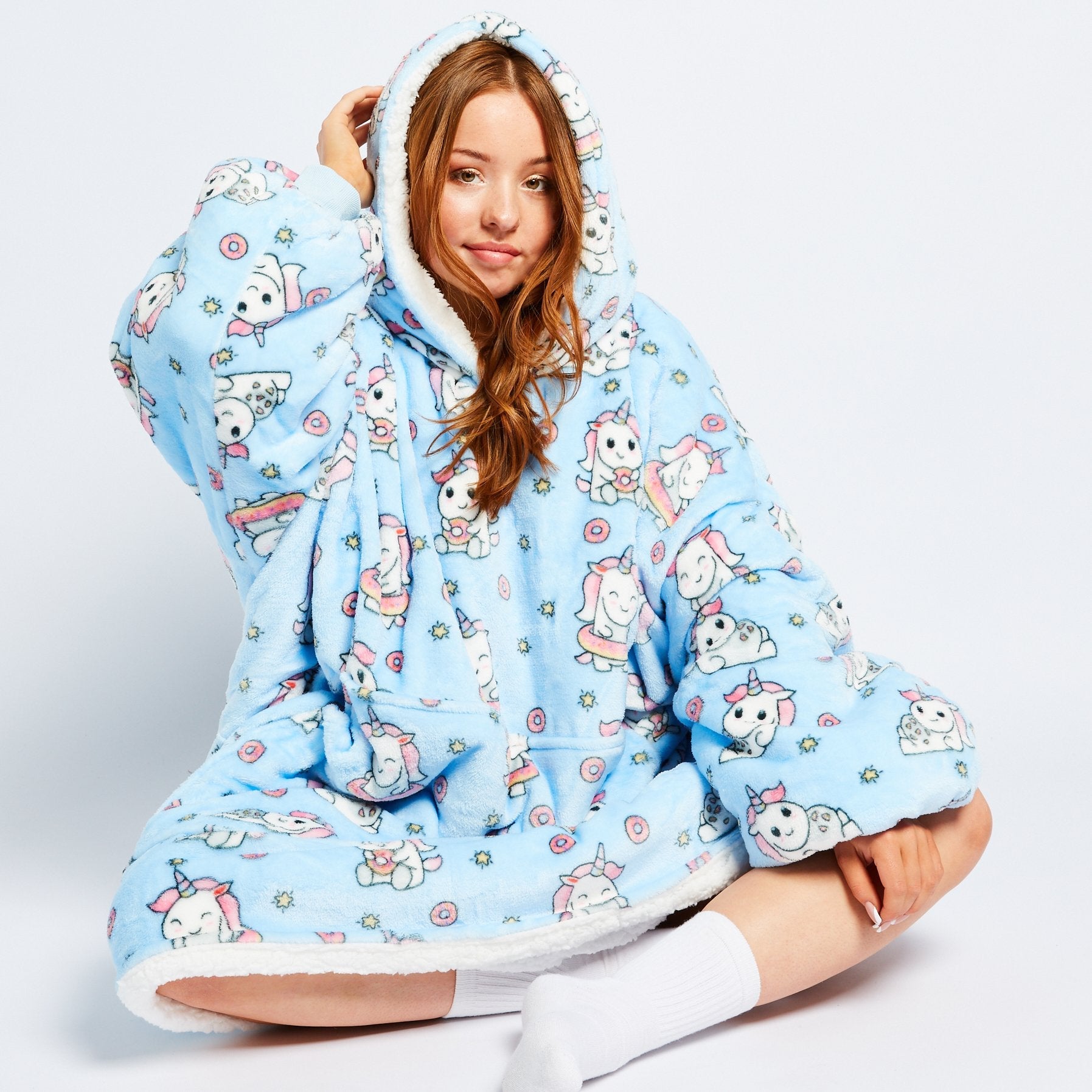 Young woman sitting cross-legged wearing a blue oversized hoodie blanket with white lining and unicorn patterns all over it.