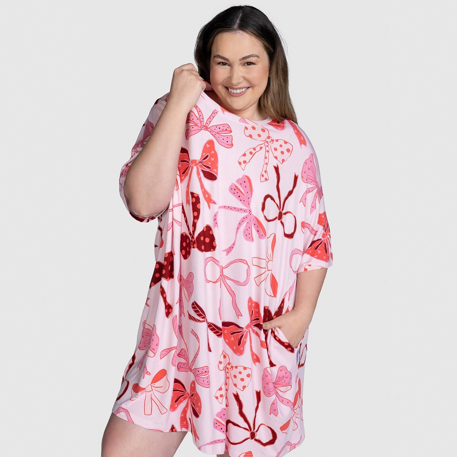 Smiling woman wearing a pink dress with various red and pink bow patterns, holding the dress near her shoulder and with one hand in a pocket