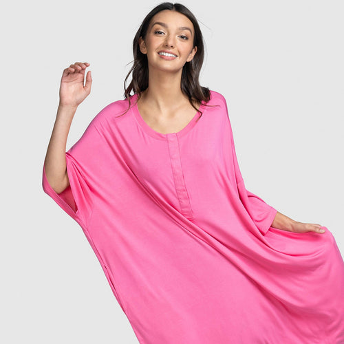 Woman smiling wearing a loose bright pink dress with wide sleeves and a round neckline, posing with one arm raised and the other holding the dress.