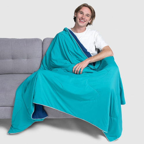 Smiling young man sitting on a gray couch wrapped in a turquoise and navy blue blanket