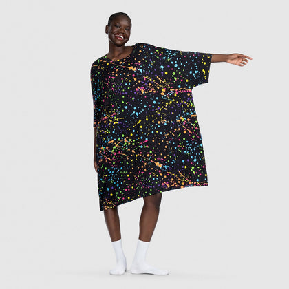 Smiling woman wearing a loose black dress with colorful paint splatter pattern, standing with one arm extended.