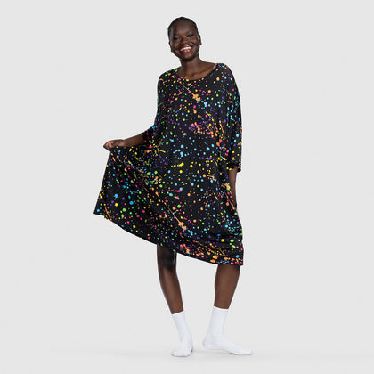 Smiling woman wearing a black dress with colorful paint splatter pattern, holding the dress out to the side.