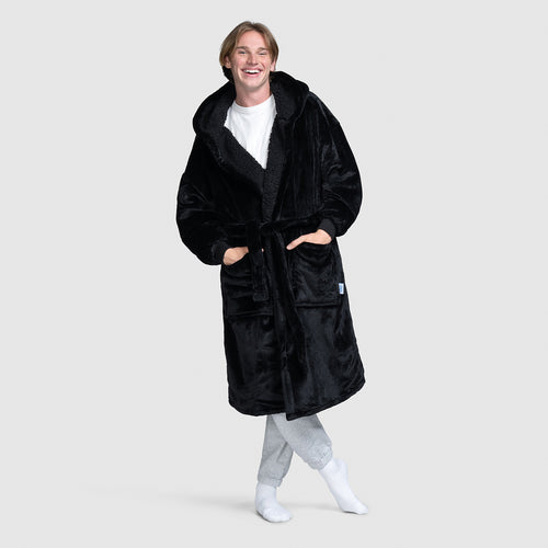 Smiling person wearing a long black plush robe with pockets and a belt, standing with hands in pockets and one foot crossed over the other.
