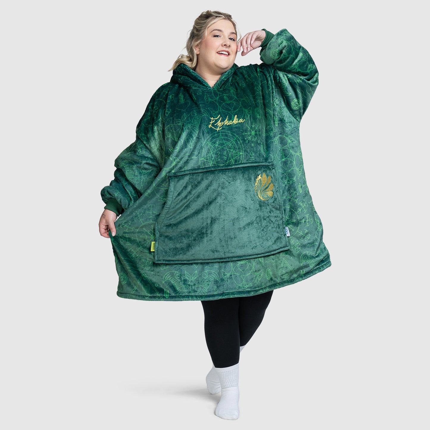 Plus-size model wearing a large green plush hoodie with 'Elphaba' embroidered on the chest and a large front pocket, standing with one arm raised.