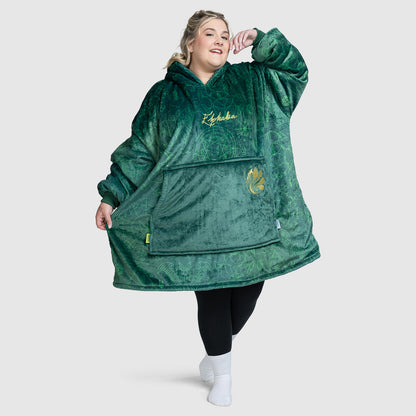 Plus-size model wearing a large green plush hoodie with 'Elphaba' embroidered on the chest and a large front pocket, standing with one arm raised.